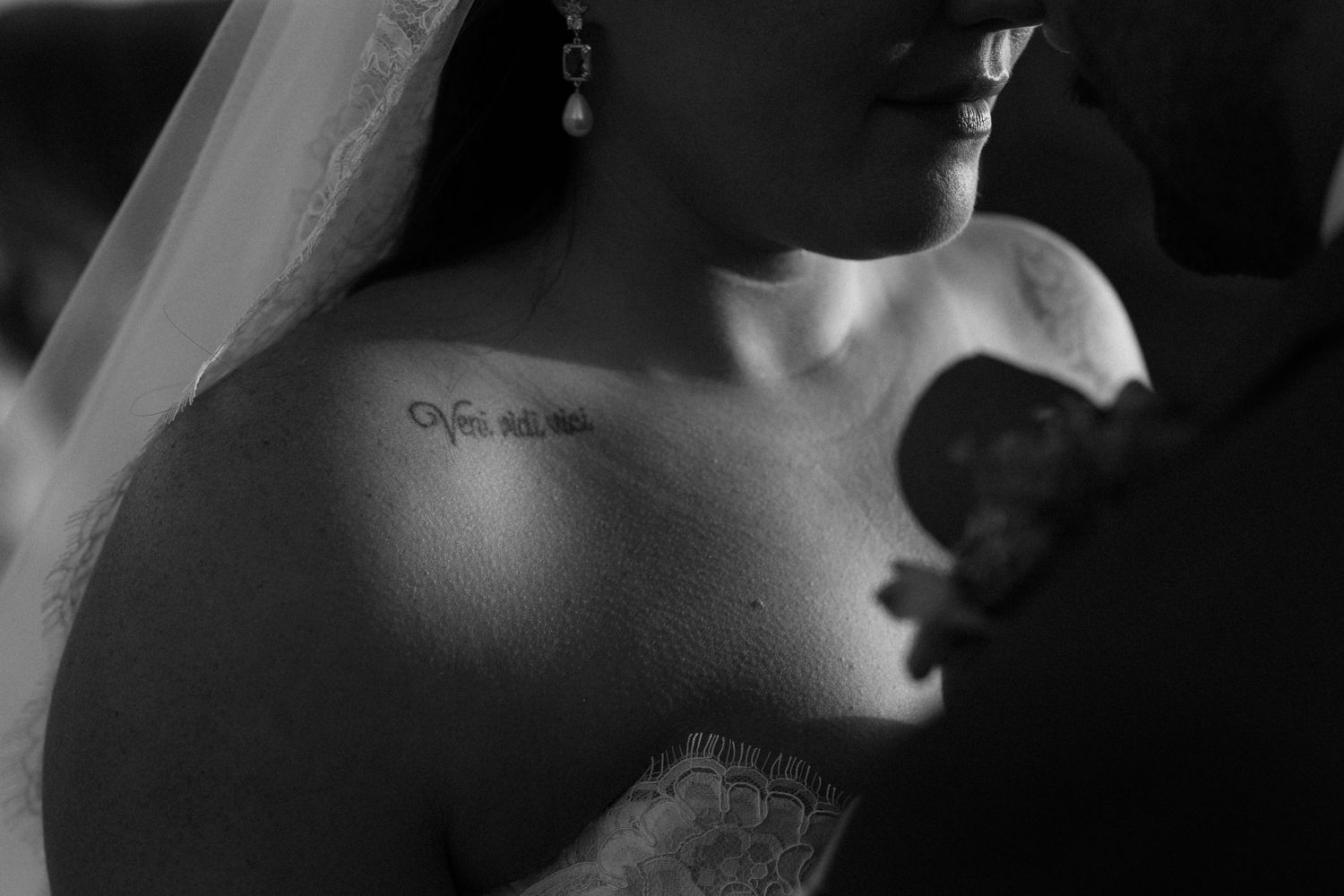 Black and white wedding photo of bride in lace dress with veil and tattoo on collarbone.