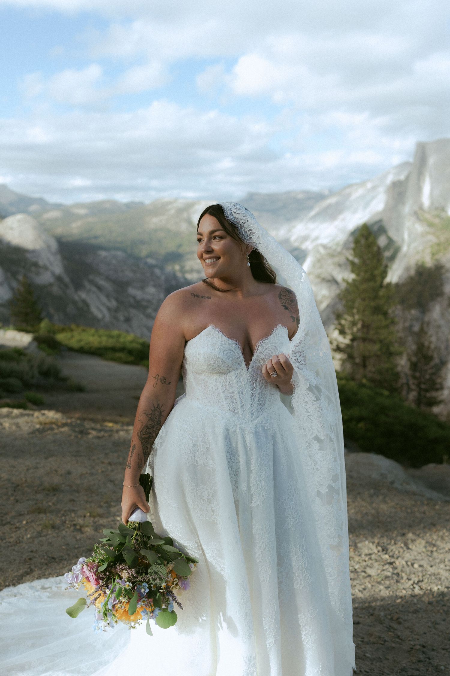 Smiling bride in strapless lace gown holding wildflower bouquet with Yosemite mountains behind her.