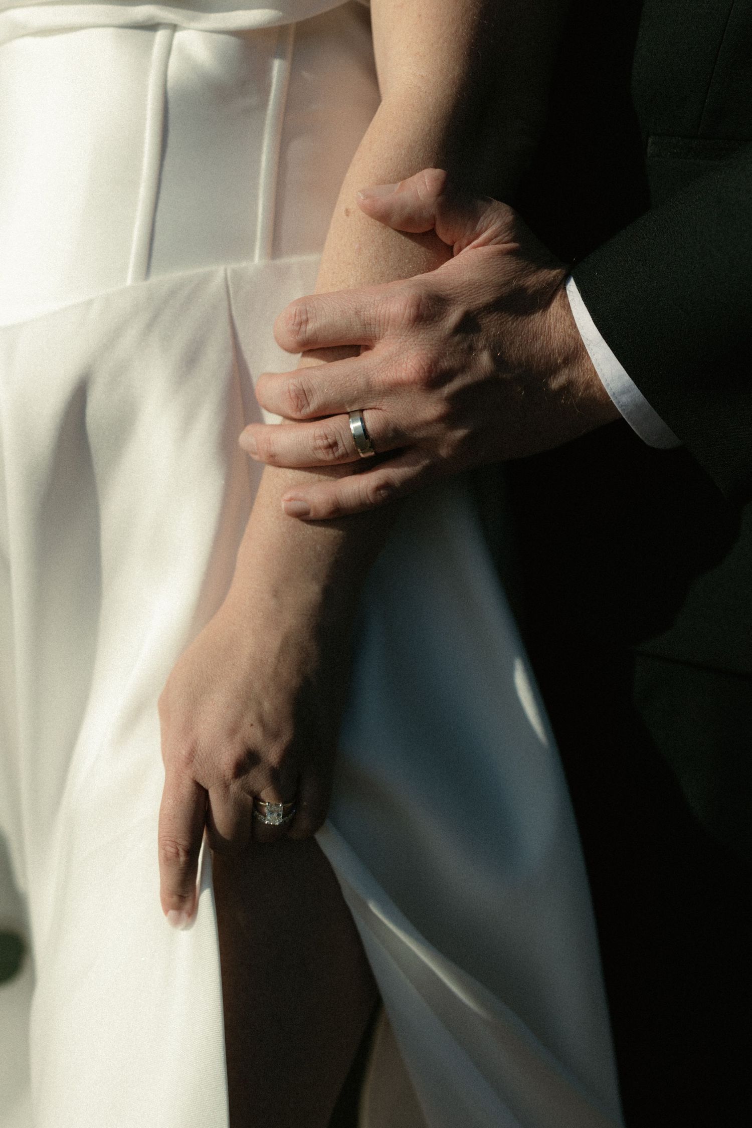 Close-up of newlywed couple holding hands, showcasing wedding and engagement rings against a white bridal gown.