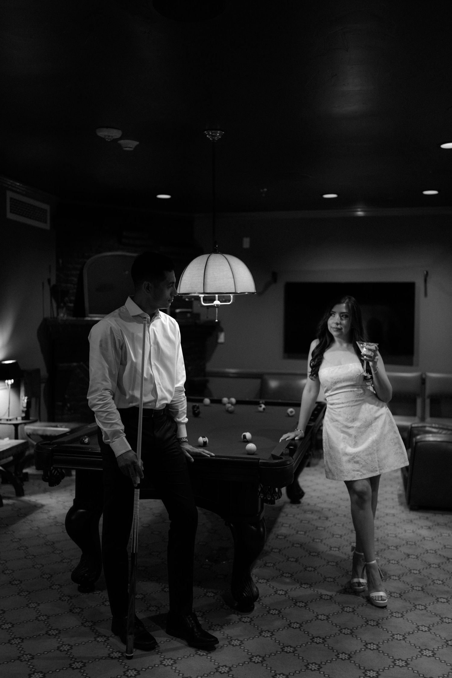 Black and white photo of a couple near a pool table in a dimly lit game room, man holding cue stick, woman in white dress.