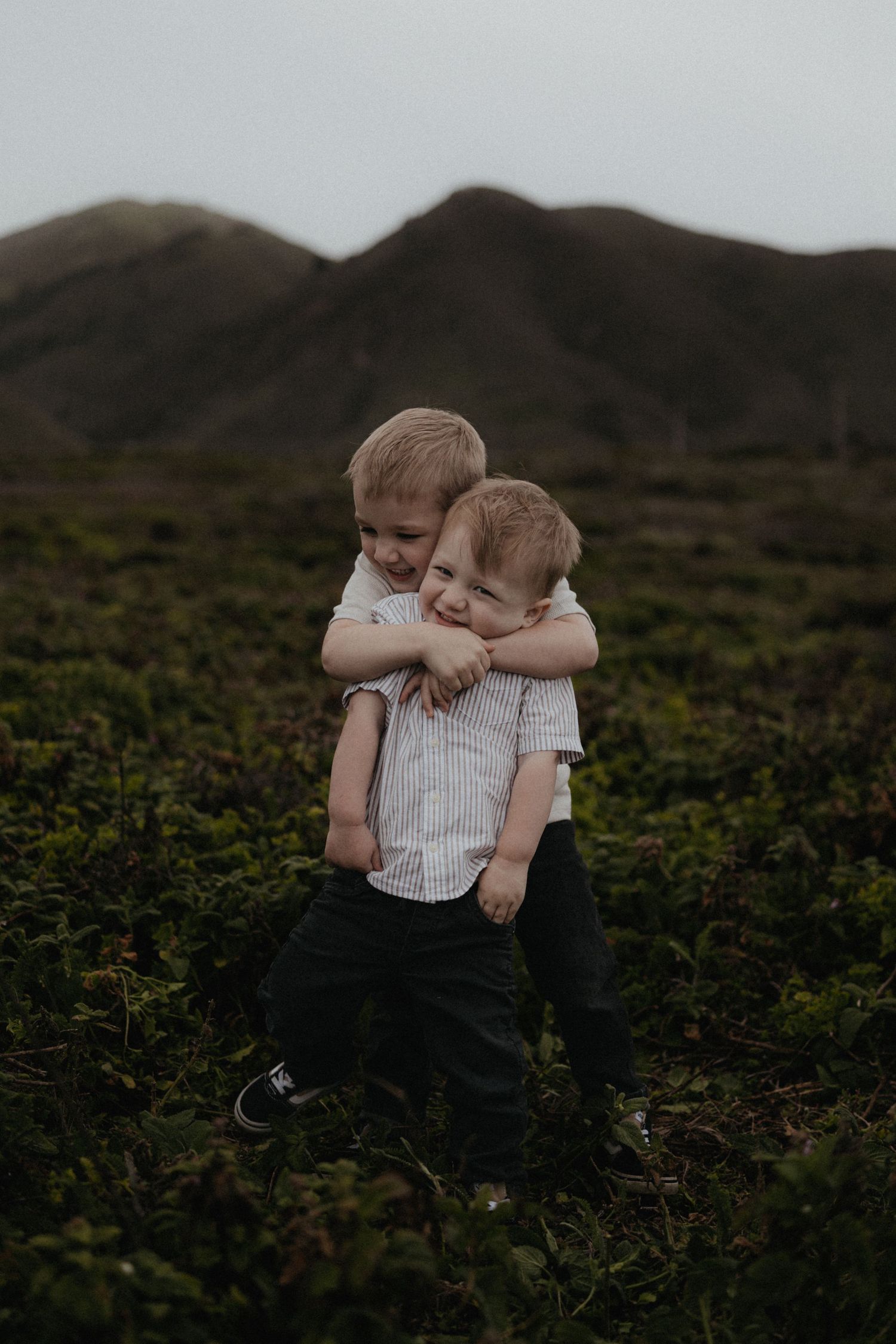 Two young boys hugging in a green field with mountains in the background.