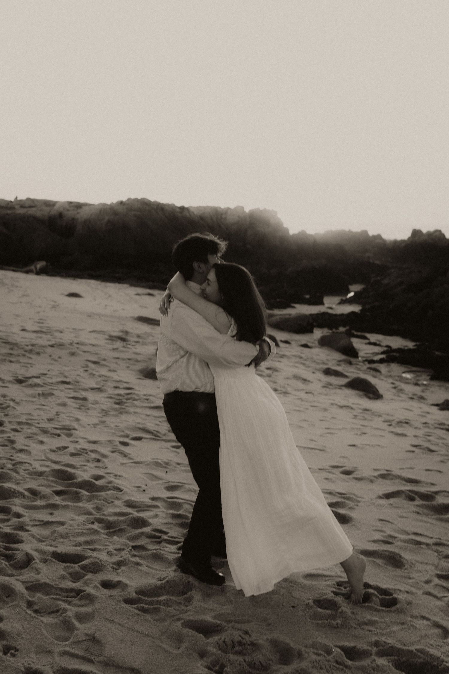 Black and white photo of a couple embracing romantically on a sandy beach with rocky cliffs in the background.