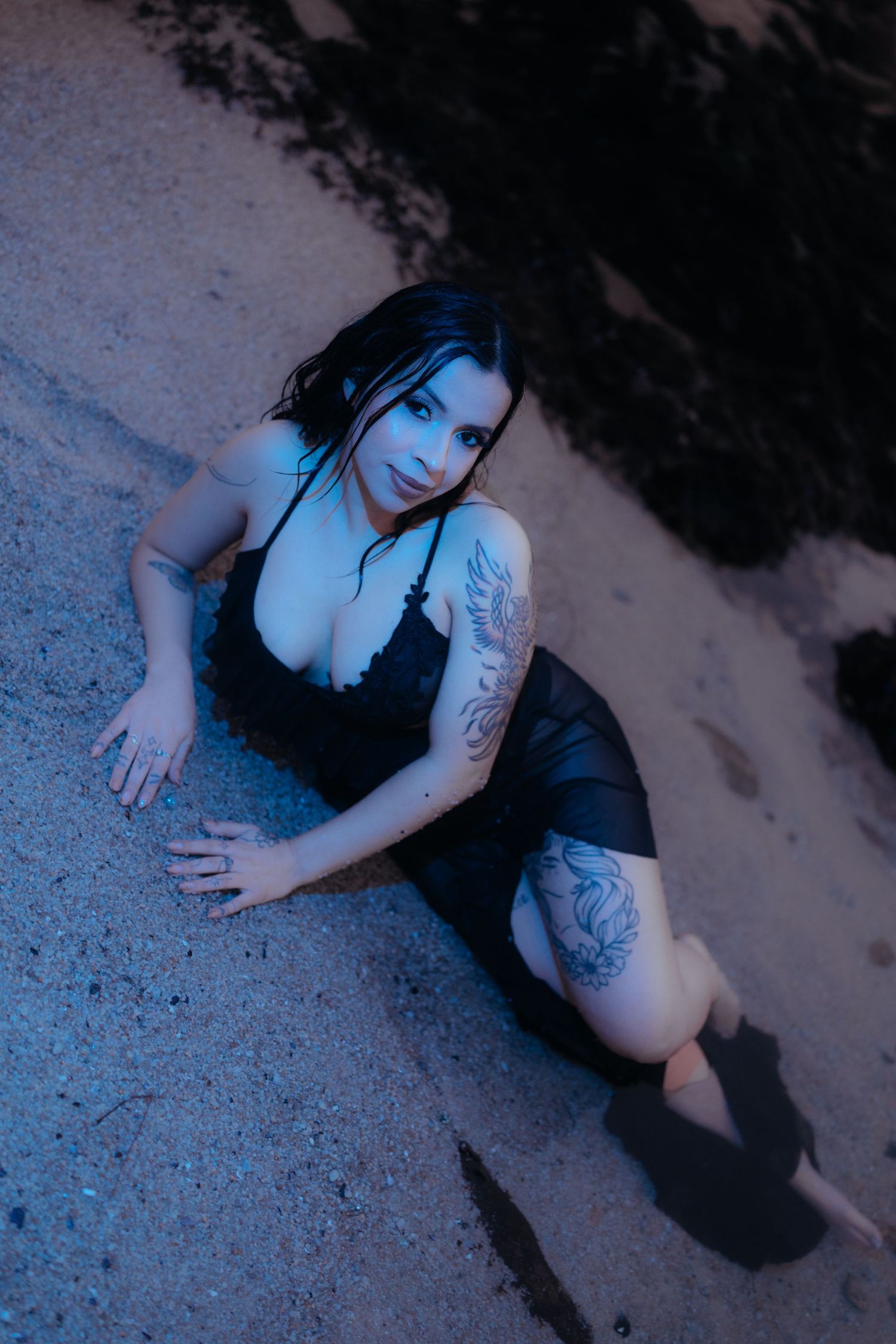 Moody portrait of a figure in black dress sitting on rocky beach at dusk.