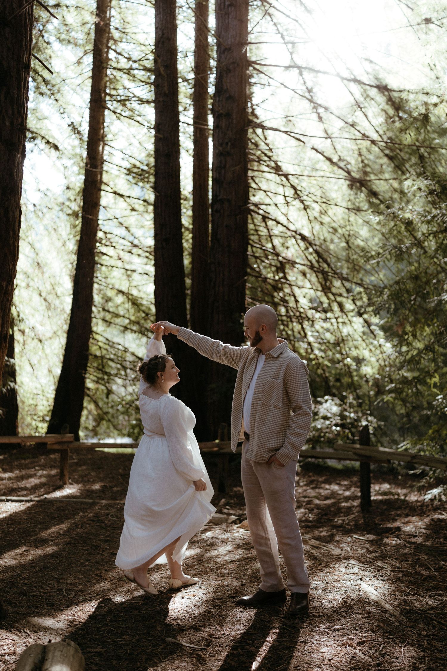 A couple in white and beige dance together in a magical forest setting with streaming sunlight.