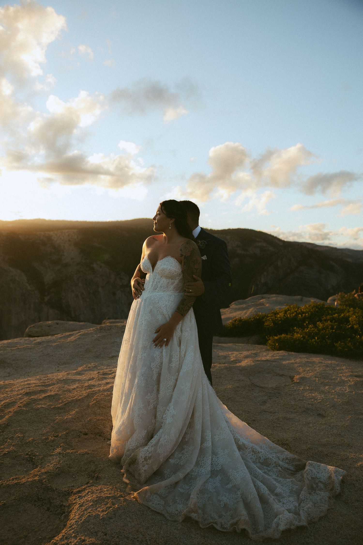 Bride and groom embracing on rocky mountaintop at golden hour with sweeping lace wedding gown train.