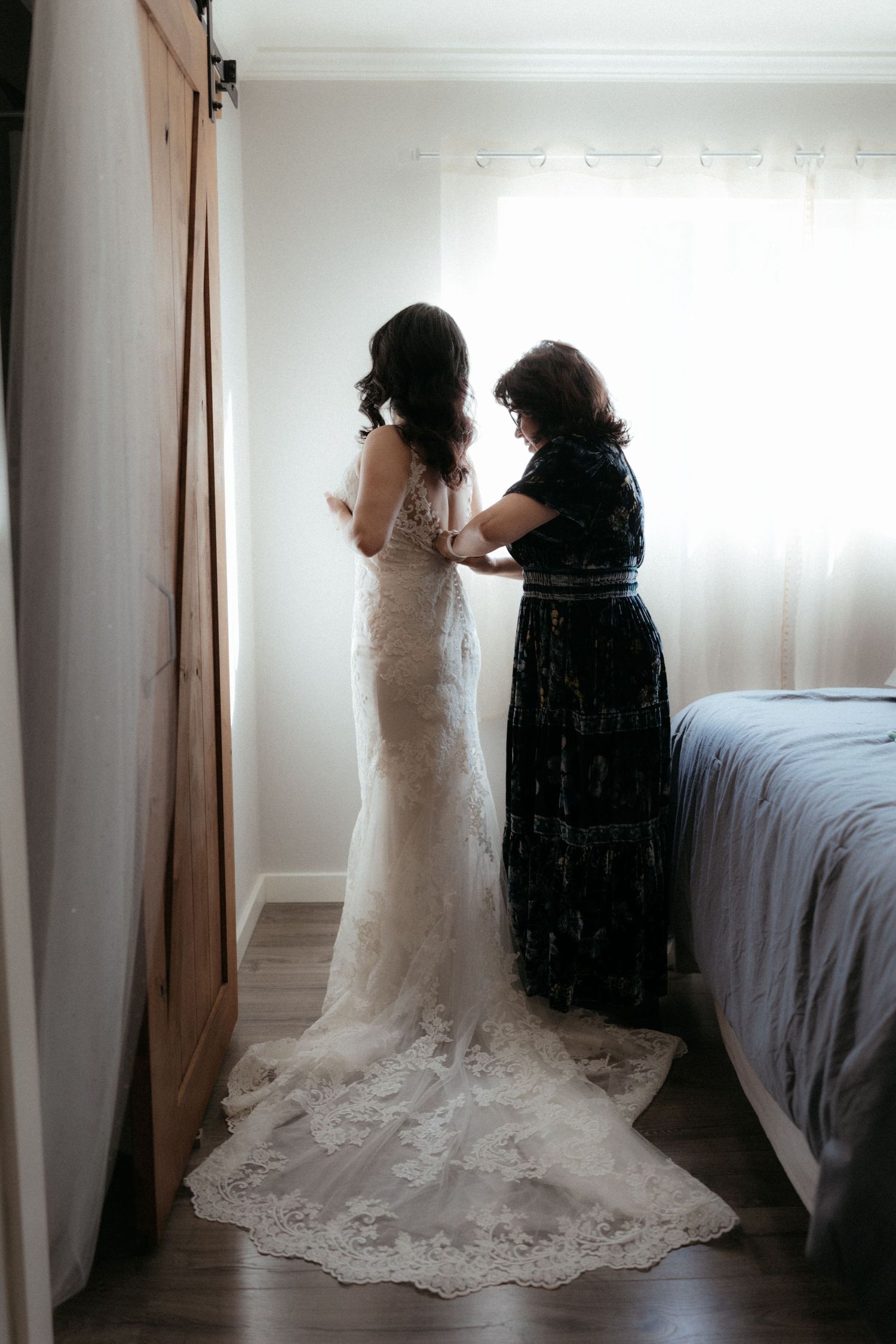 A bride in a lace wedding dress with a long train gets help with final adjustments in a bedroom.