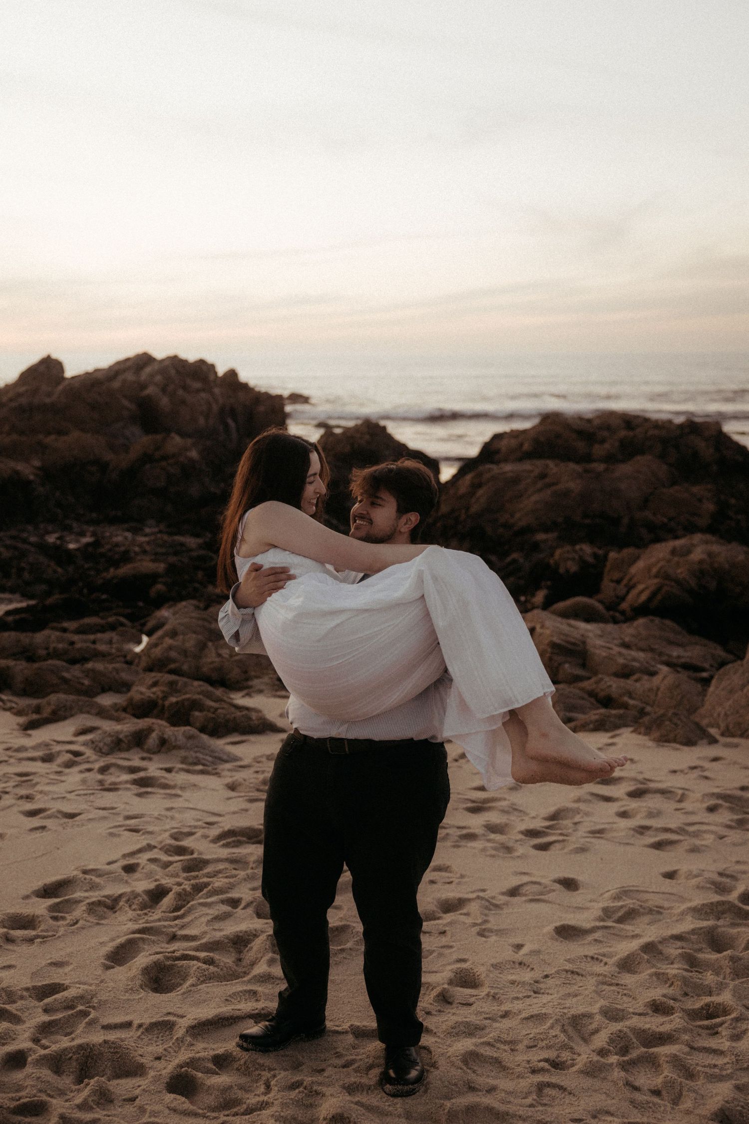 Groom carrying bride in white dress on sandy beach at sunset with rocky coastline backdrop.
