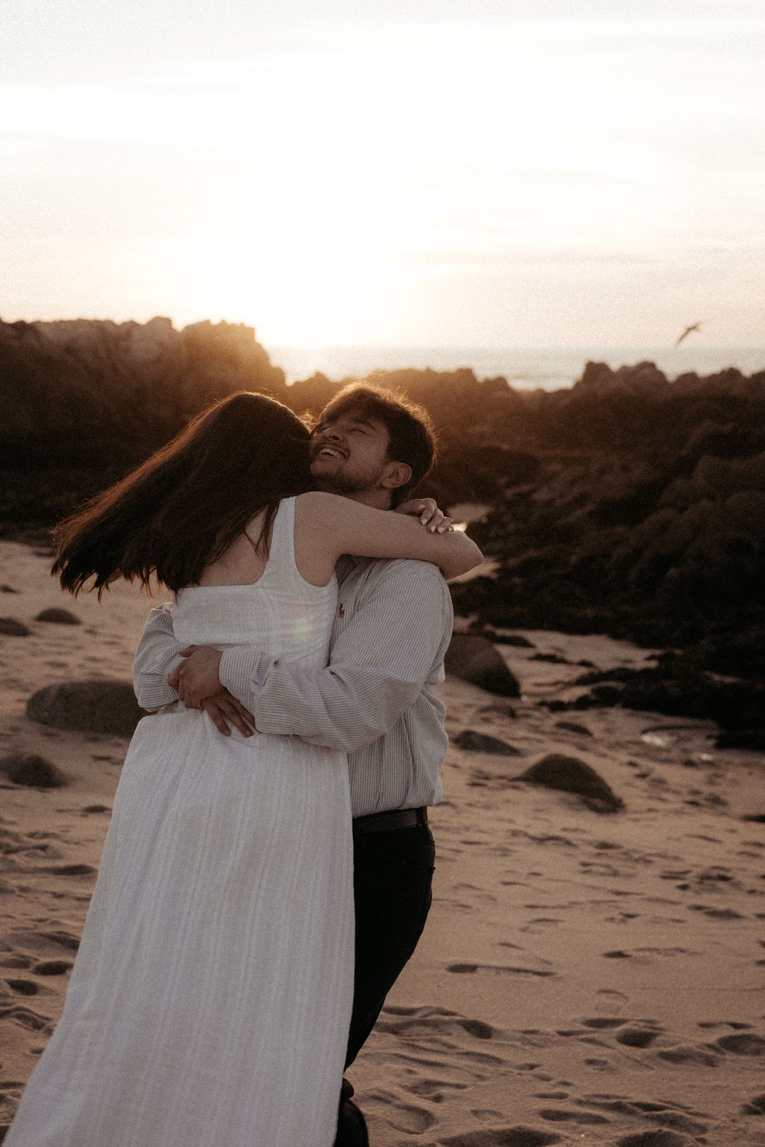 Couple embracing on a sandy beach at golden sunset with rocky cliffs in background.