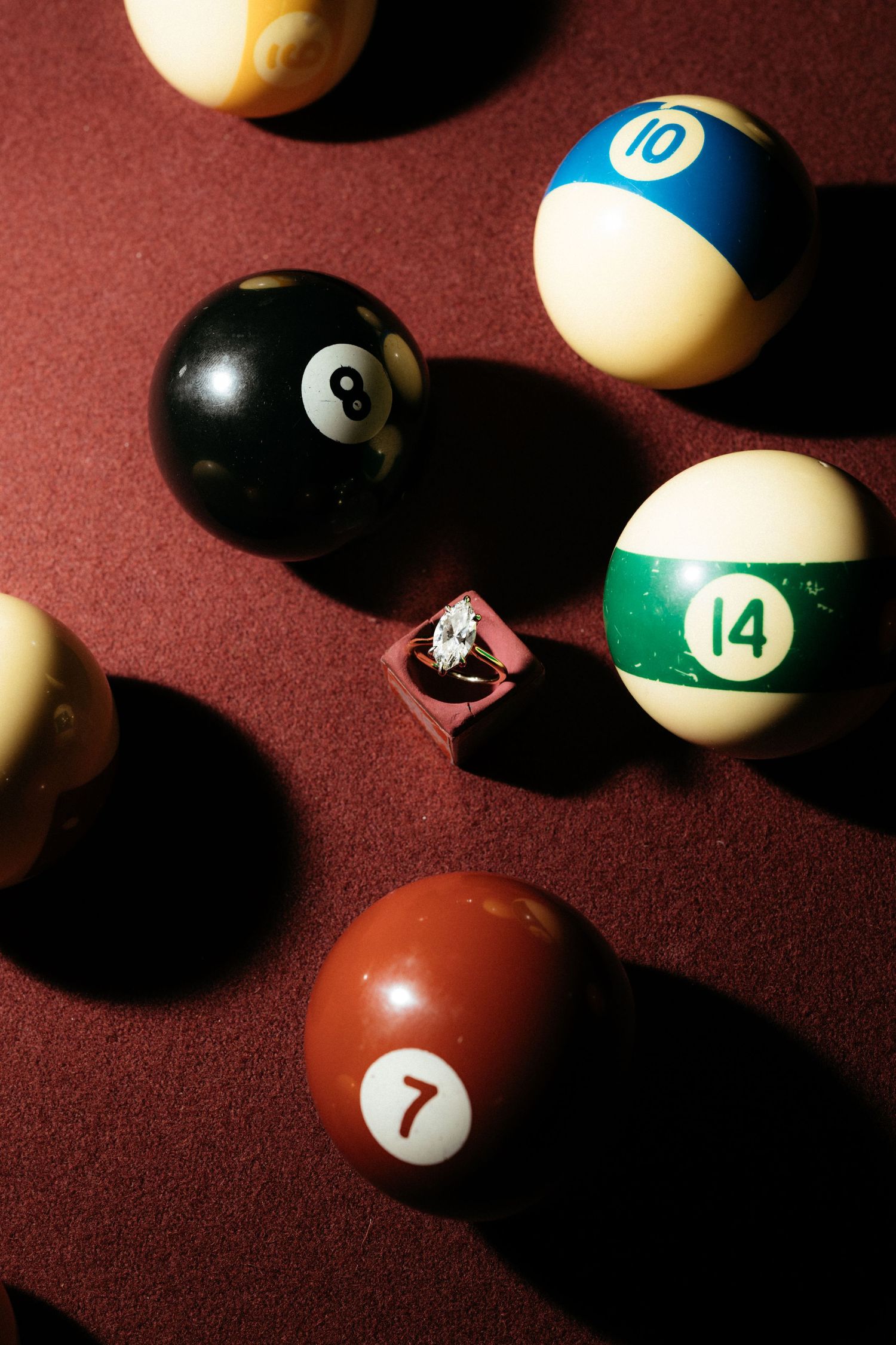 An engagement ring in a pink box surrounded by billiard balls including the 8, 10, 14, and 7 on a red pool table.