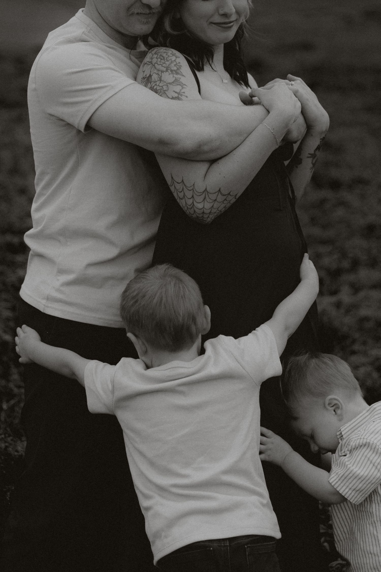Black and white family photo of parents embracing while two young boys cling to them outdoors.
