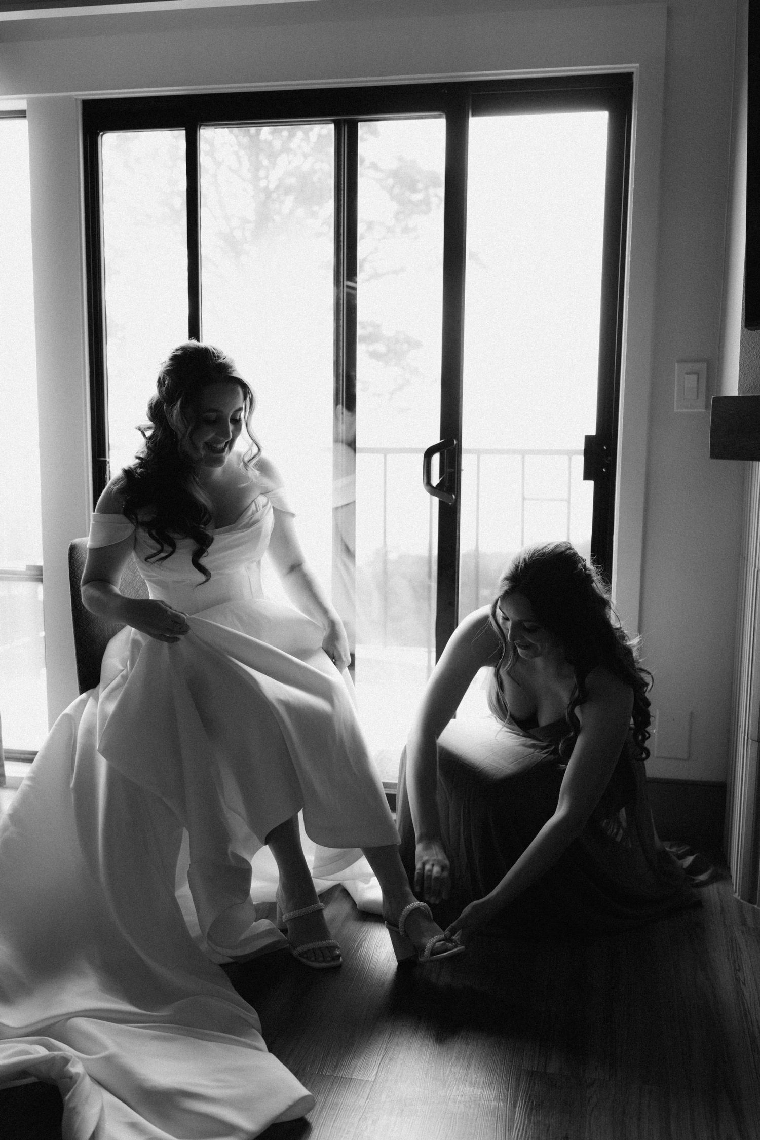Black and white photo of a bride in a white gown sitting as a bridesmaid helps fasten her wedding shoes near glass doors.