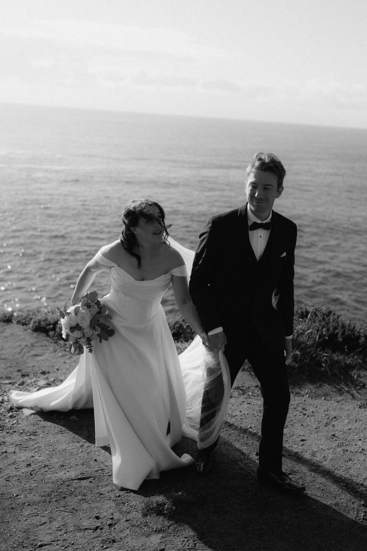 Bride and groom walk hand in hand along ocean cliffs in elegant black and white wedding photo.