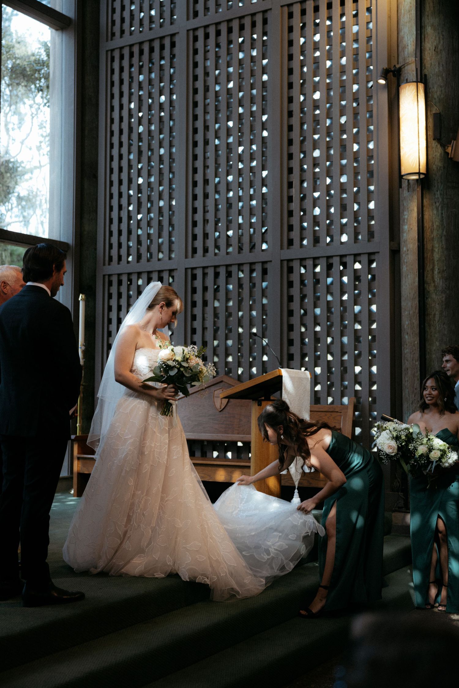 Bride at altar holding white bouquet while bridesmaid in green dress adjusts her gown during wedding ceremony.