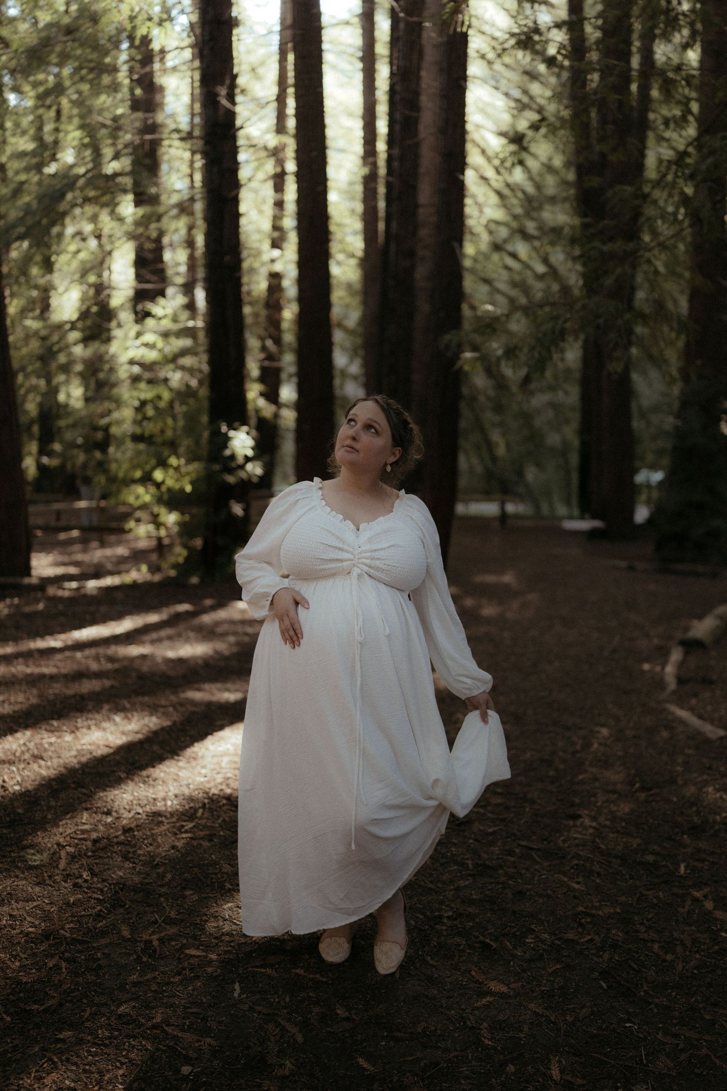 A person in a flowing white dress poses gracefully among tall forest trees in golden light.