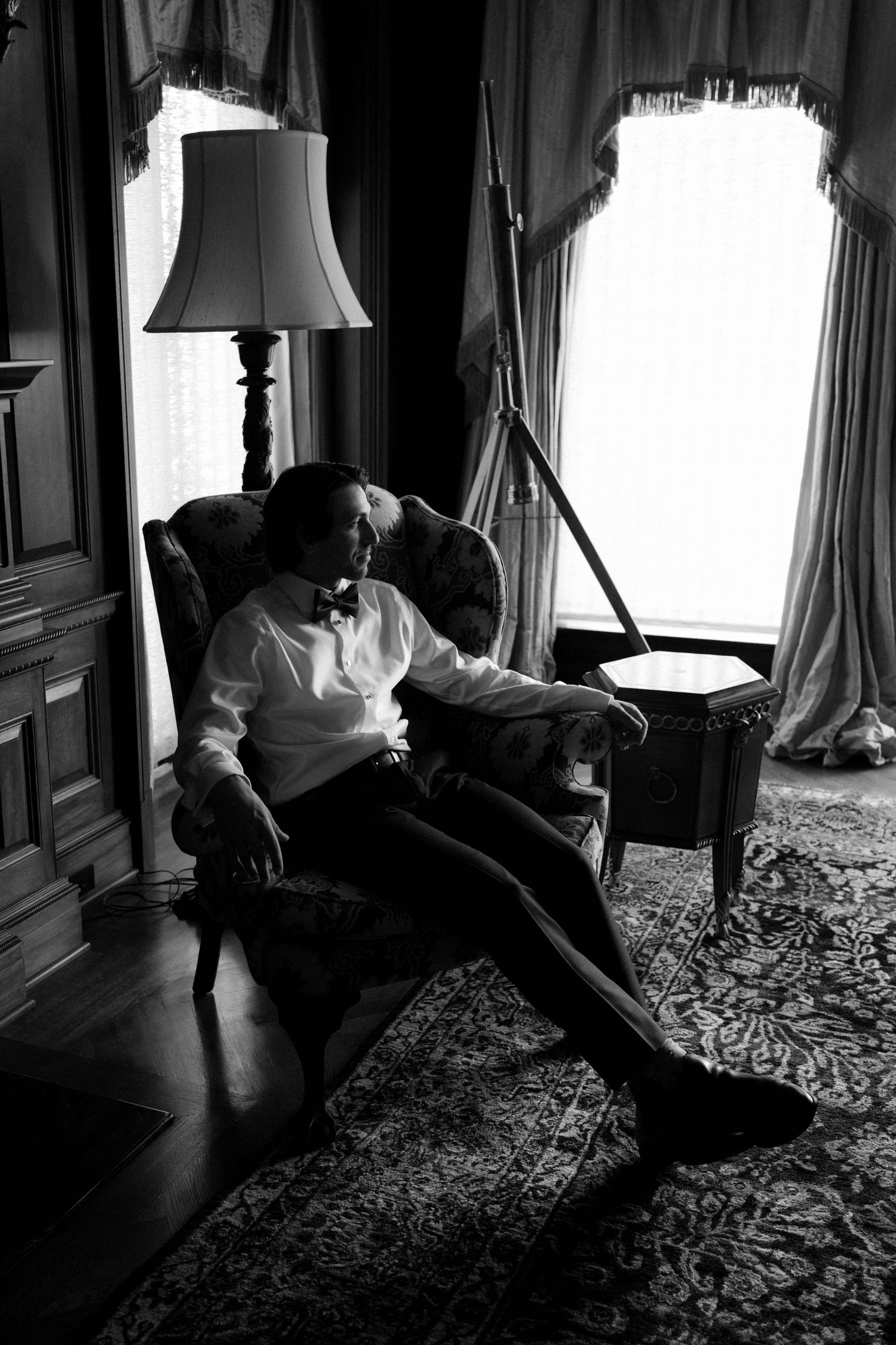 A groom in a tuxedo and bow tie sits relaxed in an armchair in an elegant room, black and white photo.