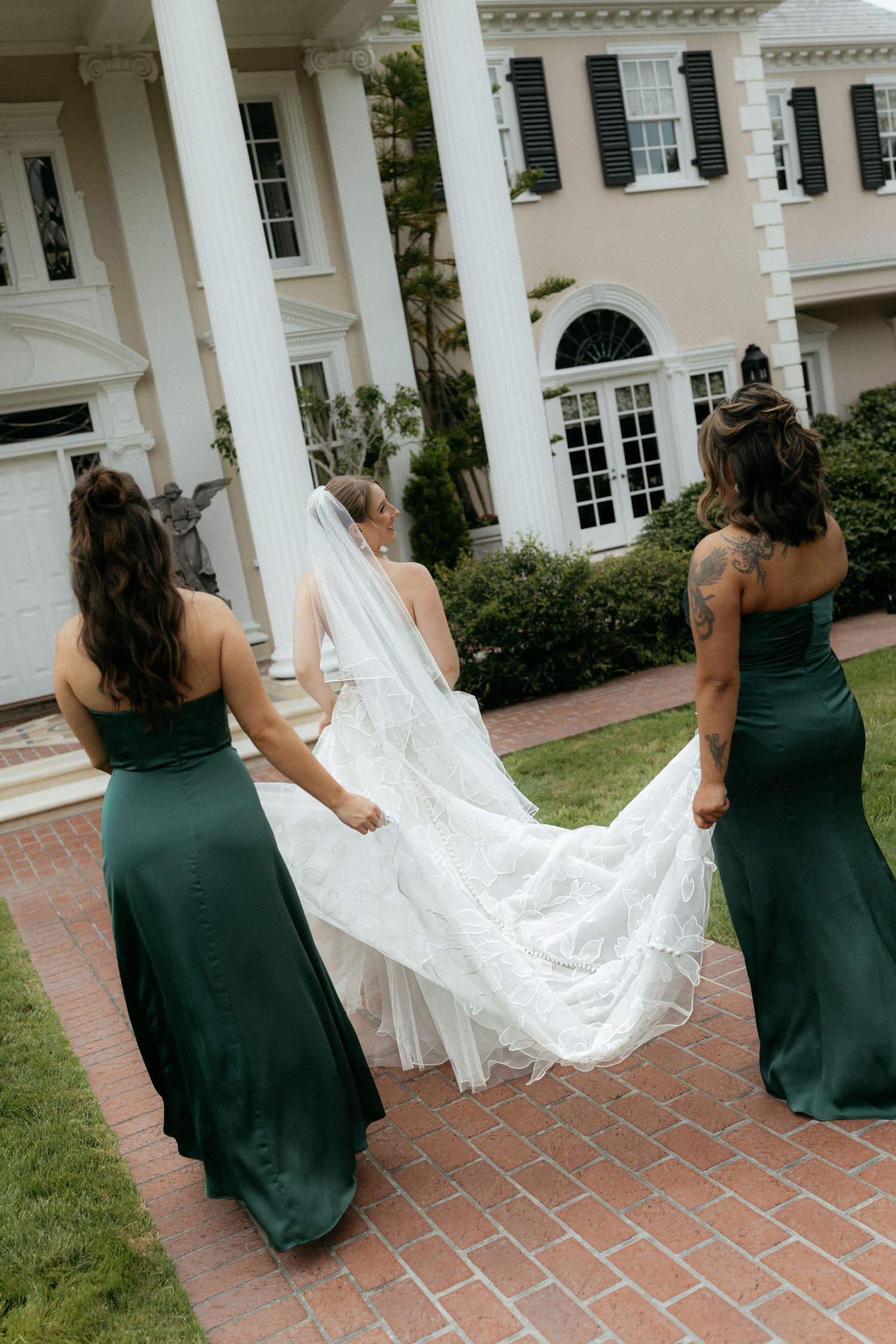 Two bridesmaids in green satin gowns carry a bride's white lace train outside an elegant estate.