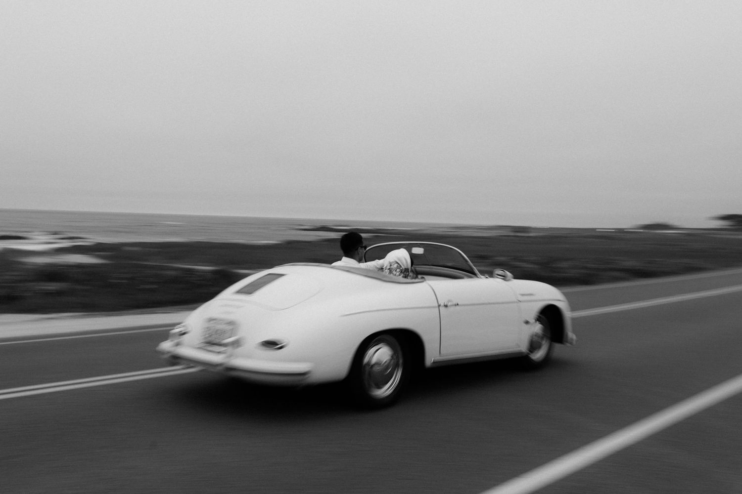 Black and white photo of a vintage white Porsche 356 Speedster cruising along a scenic ocean road.