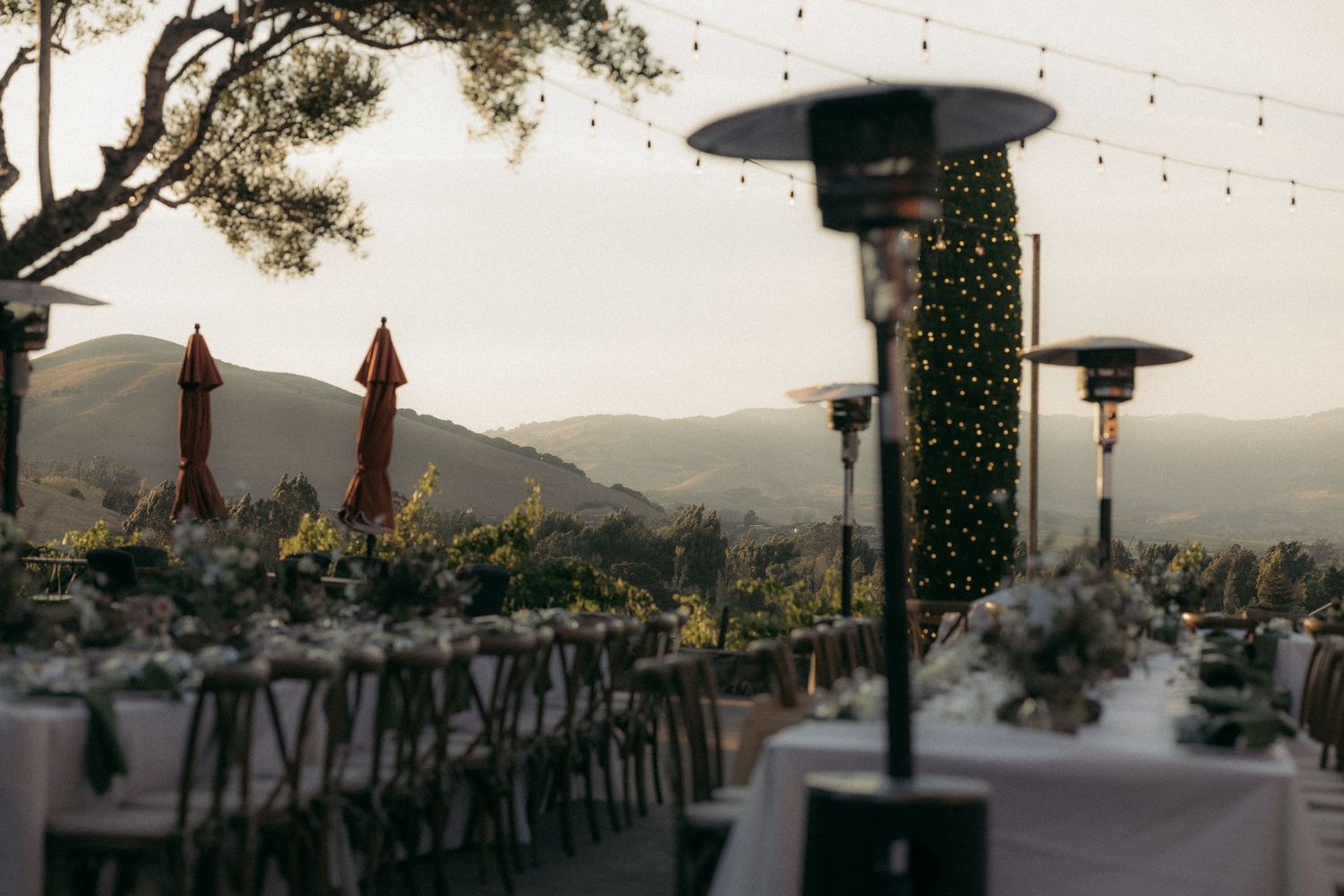 Outdoor wedding reception setup with tables and chairs overlooking mountain views at sunset with hanging patio heaters.