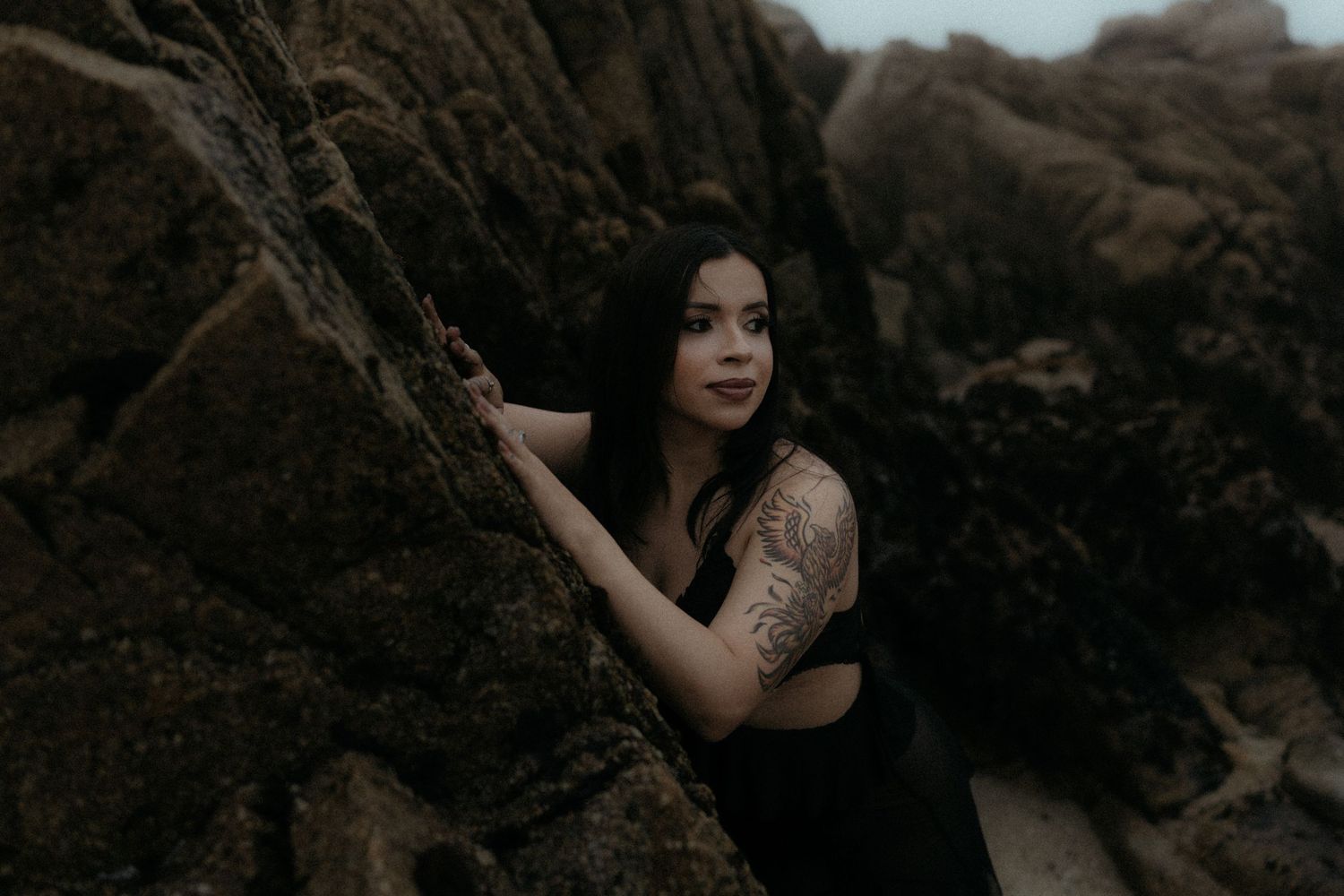 A person in a black dress poses against a dark rocky background in a moody outdoor photoshoot.