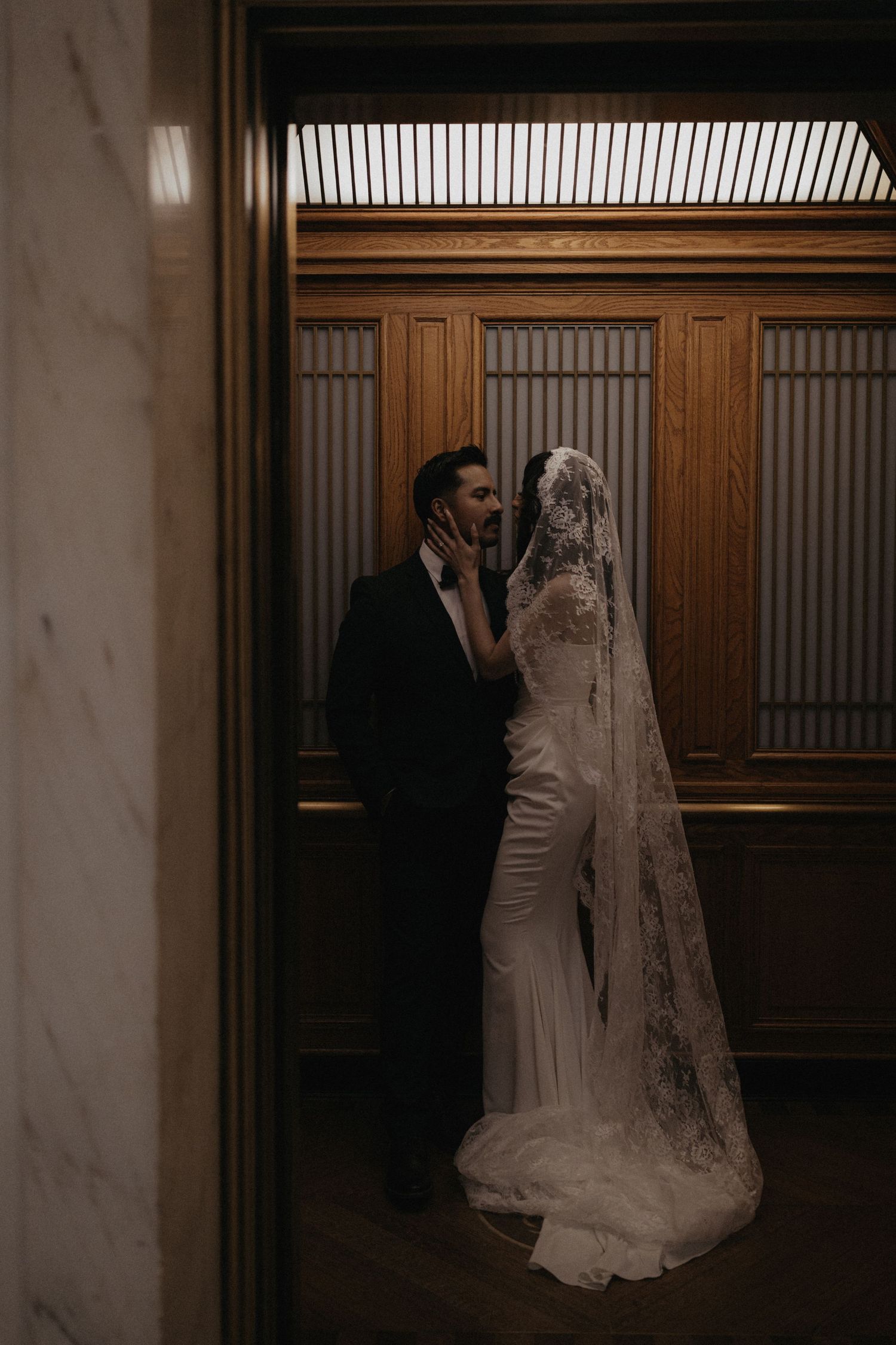 A bride in a white lace wedding gown and veil stands in a dimly lit hallway with wooden paneled walls.