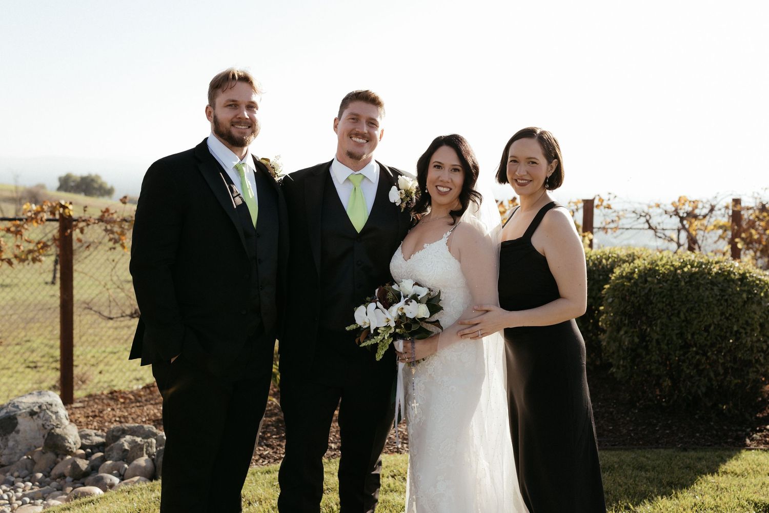 Wedding party poses outdoors in formal attire with bridesmaids in black dresses and groomsmen in black suits.