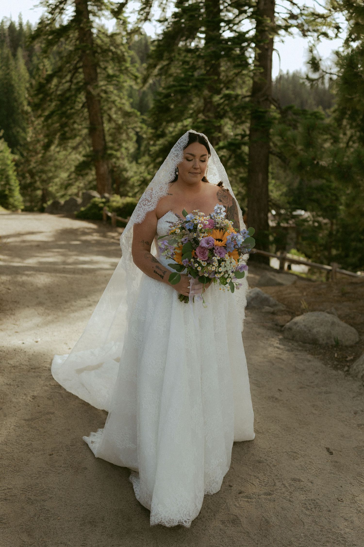 Bride in white lace gown and veil holds colorful bouquet on forest path.