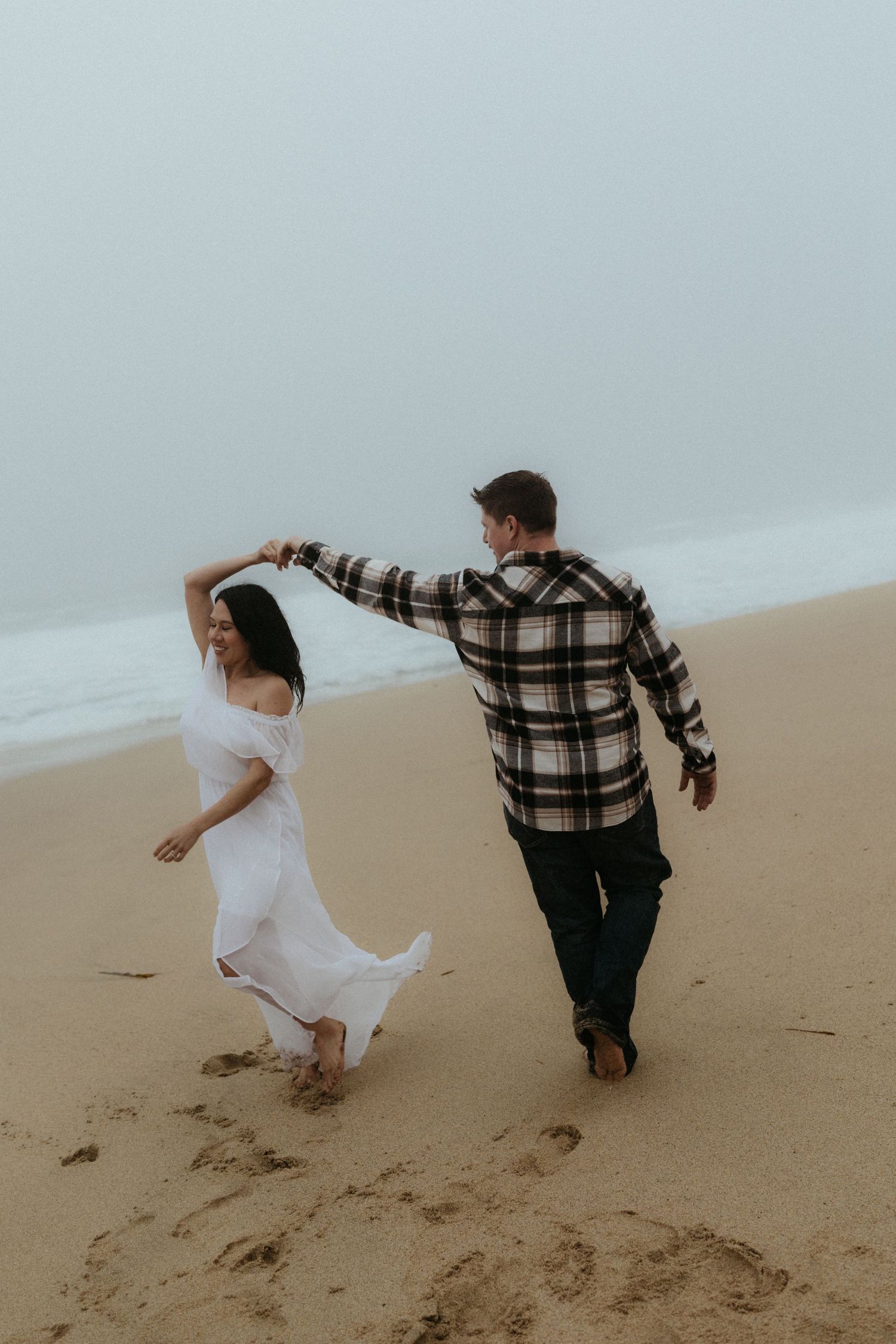 Two people run playfully along the beach shore during an overcast day.