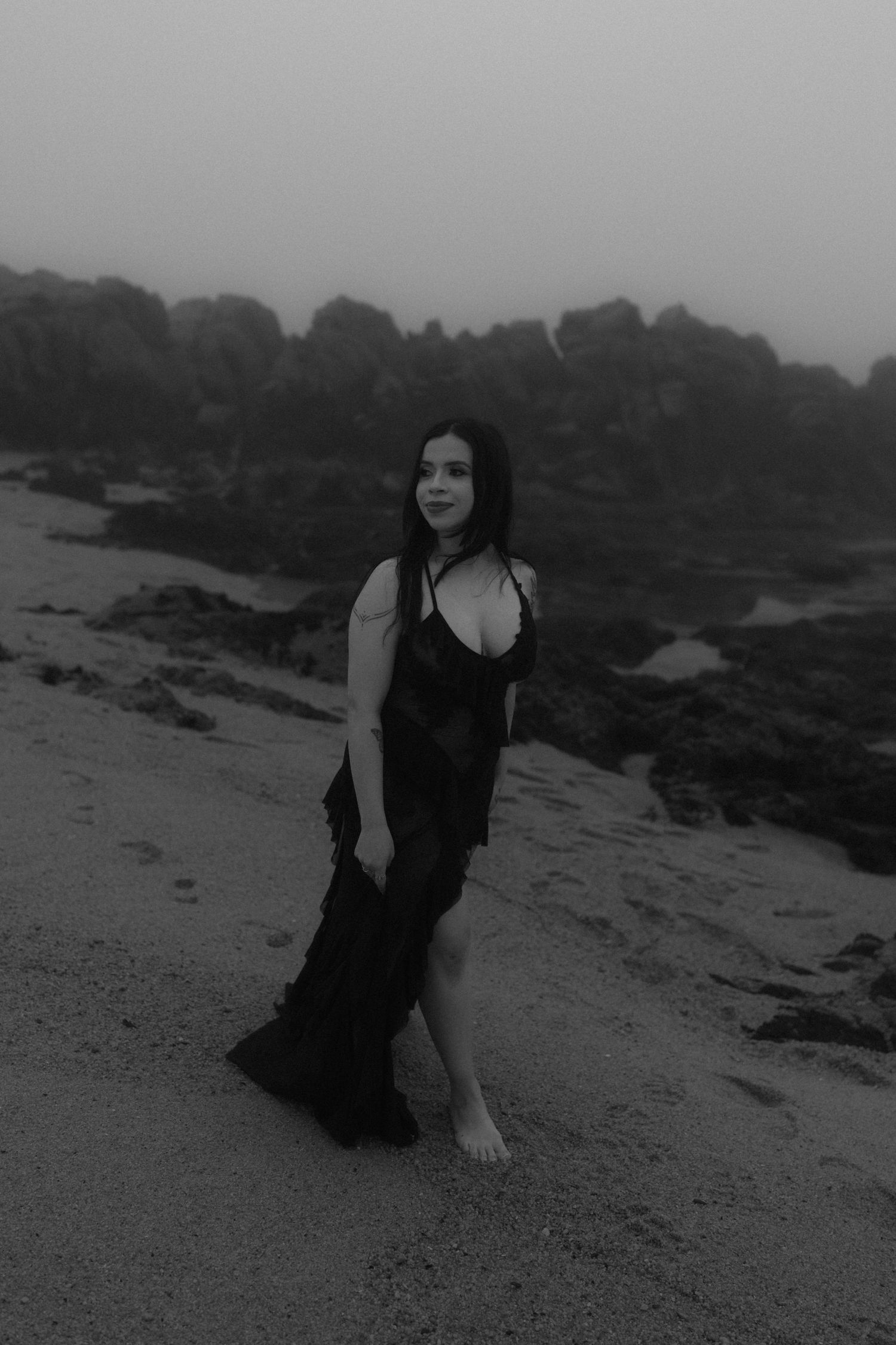 Figure in black dress poses on rocky beach in atmospheric monochrome shot.