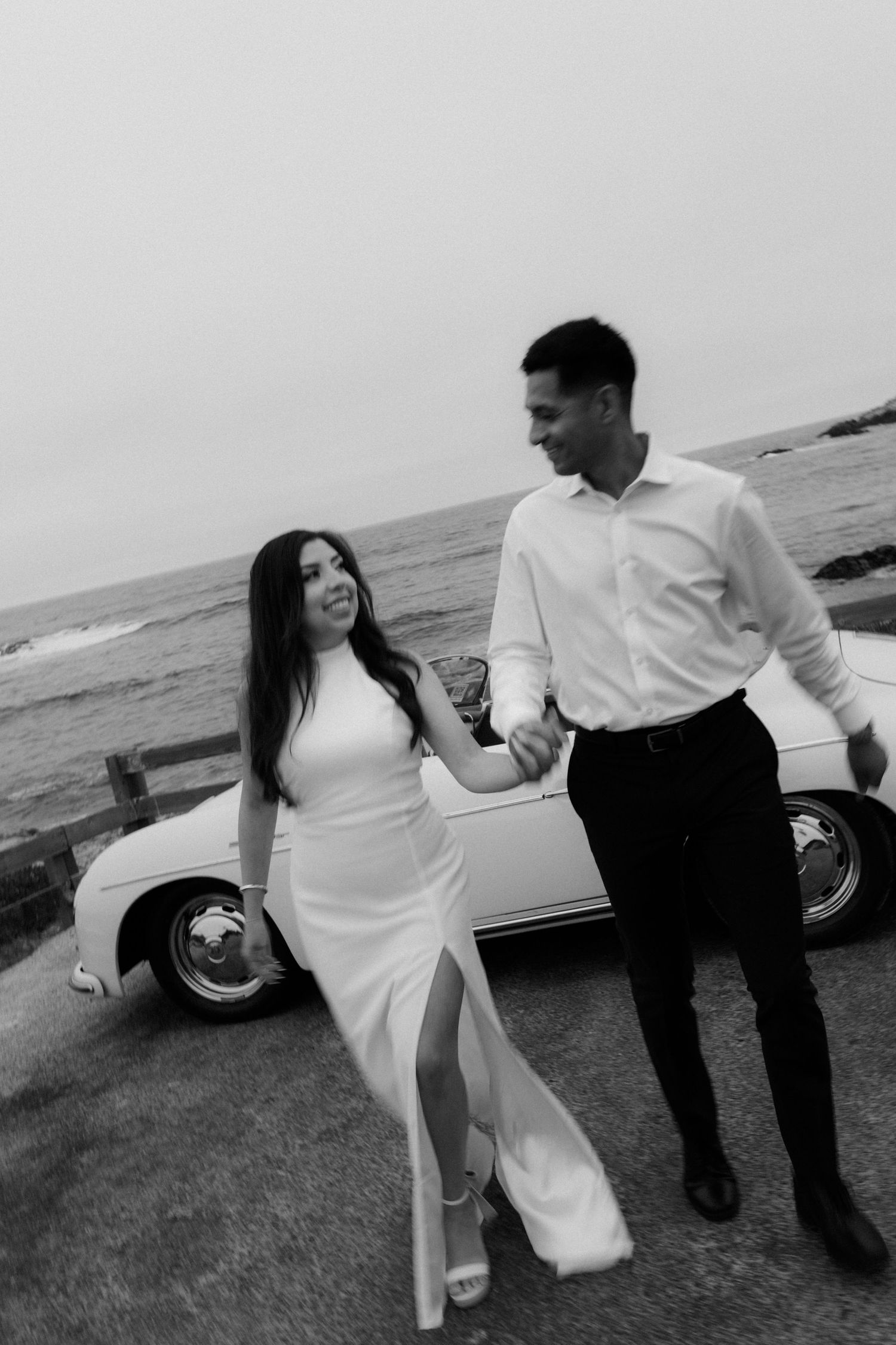 Black and white photo of a couple holding hands by a vintage car near the ocean.