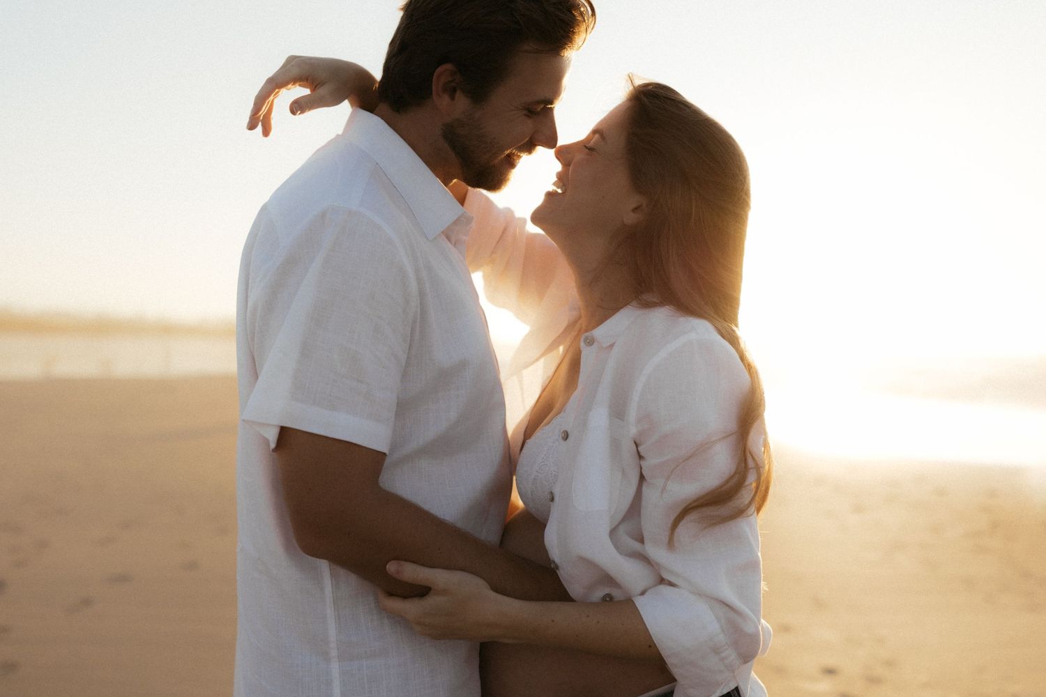 Romantic couple embracing on a sunny beach at golden hour, wearing white outfits, about to kiss.