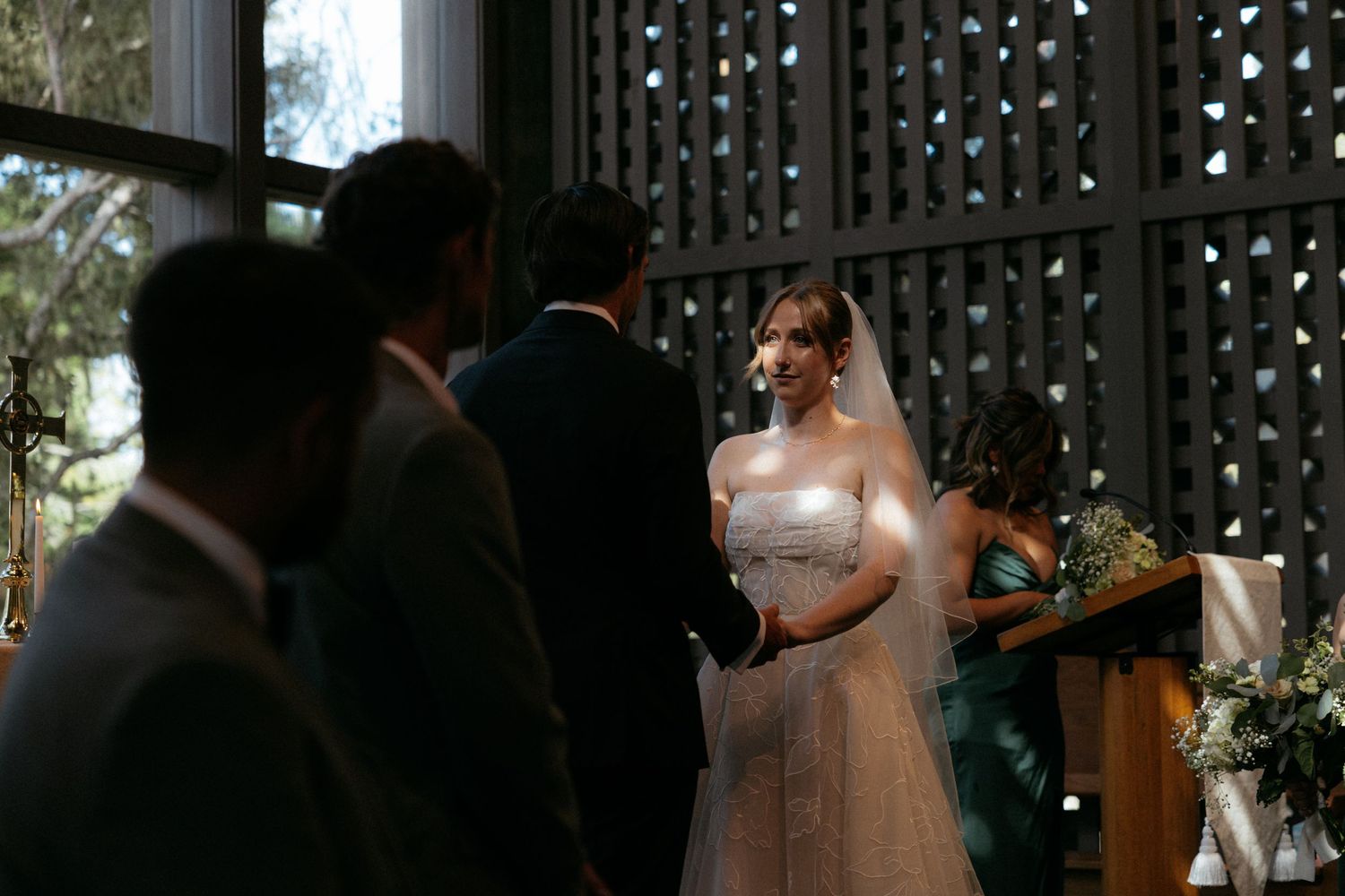 Bride smiling at groom during indoor wedding ceremony, wearing strapless white gown with veil, guests and officiant visible.