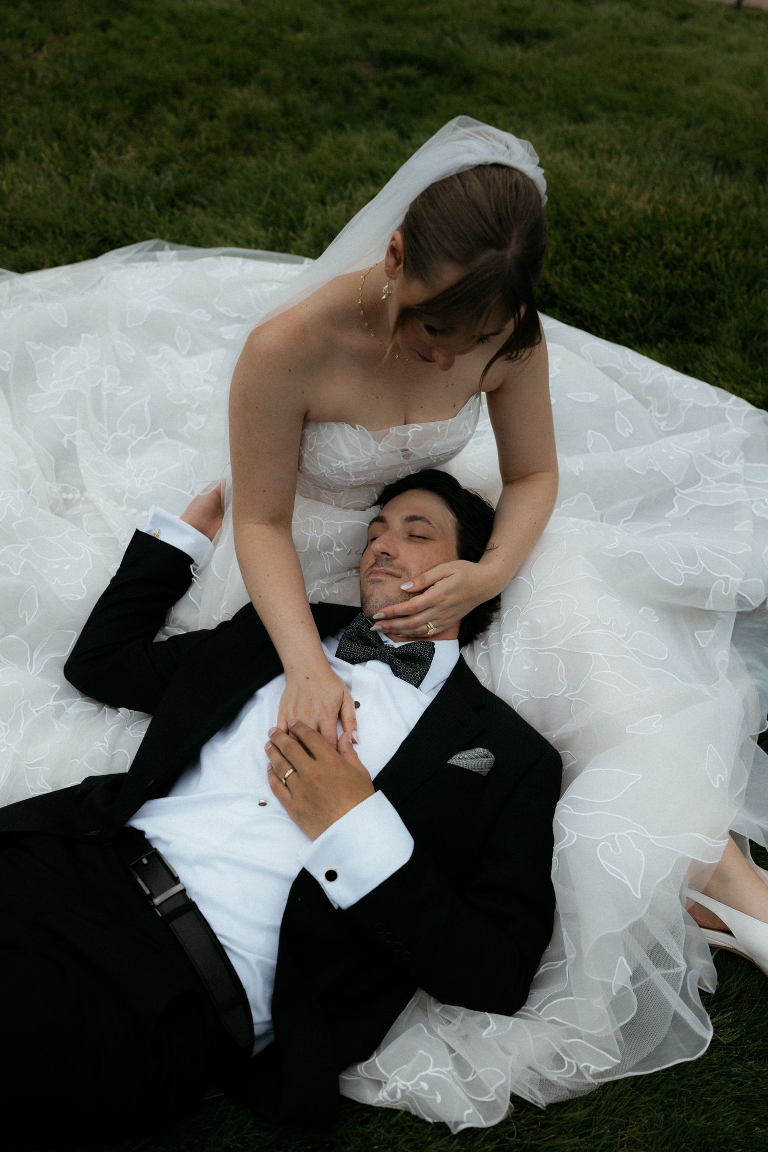 Bride in white gown sits on grass cradling groom in black tuxedo lying across her wedding dress outdoors.