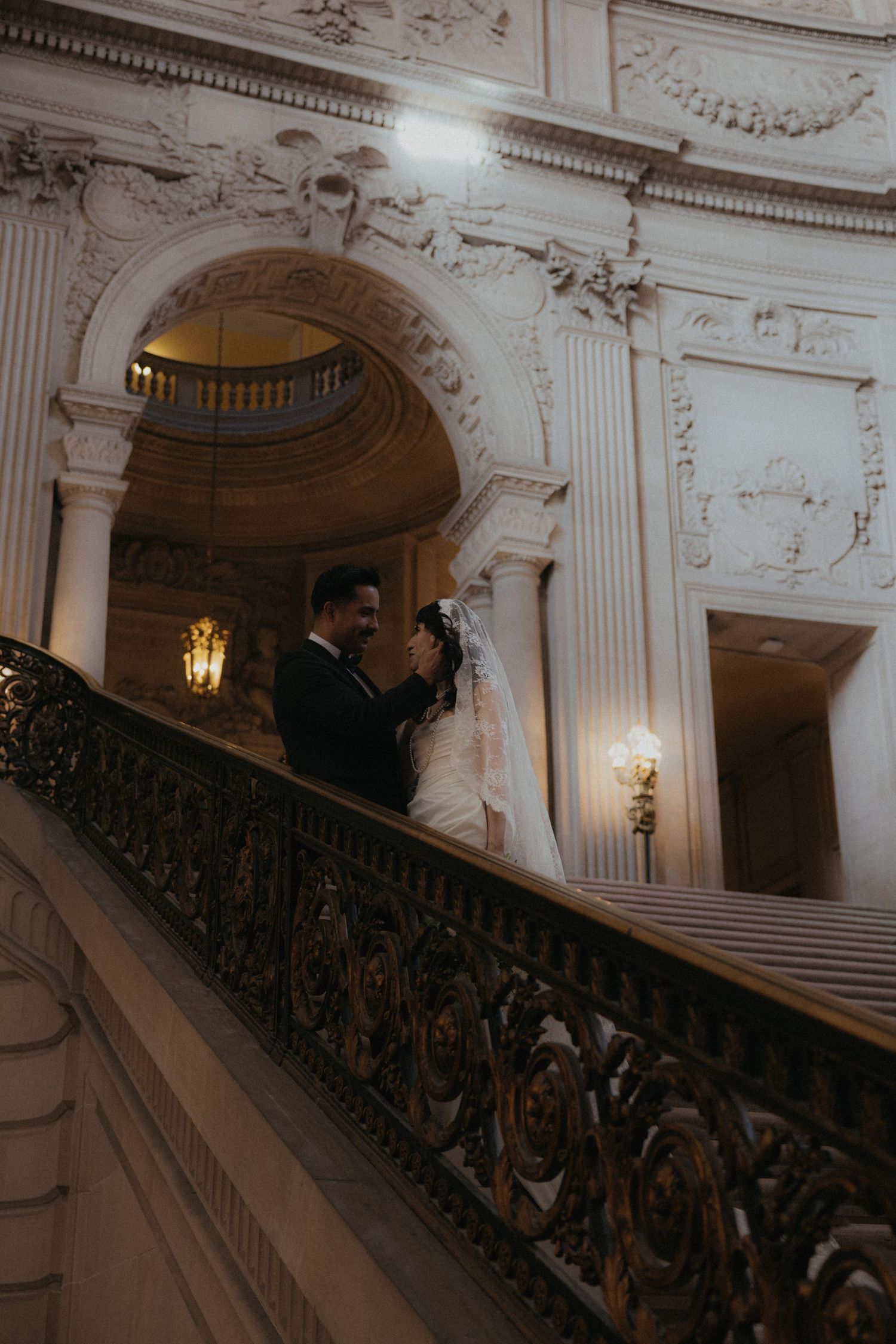 A bride and groom share a romantic moment on an ornate staircase in a grand historic building.