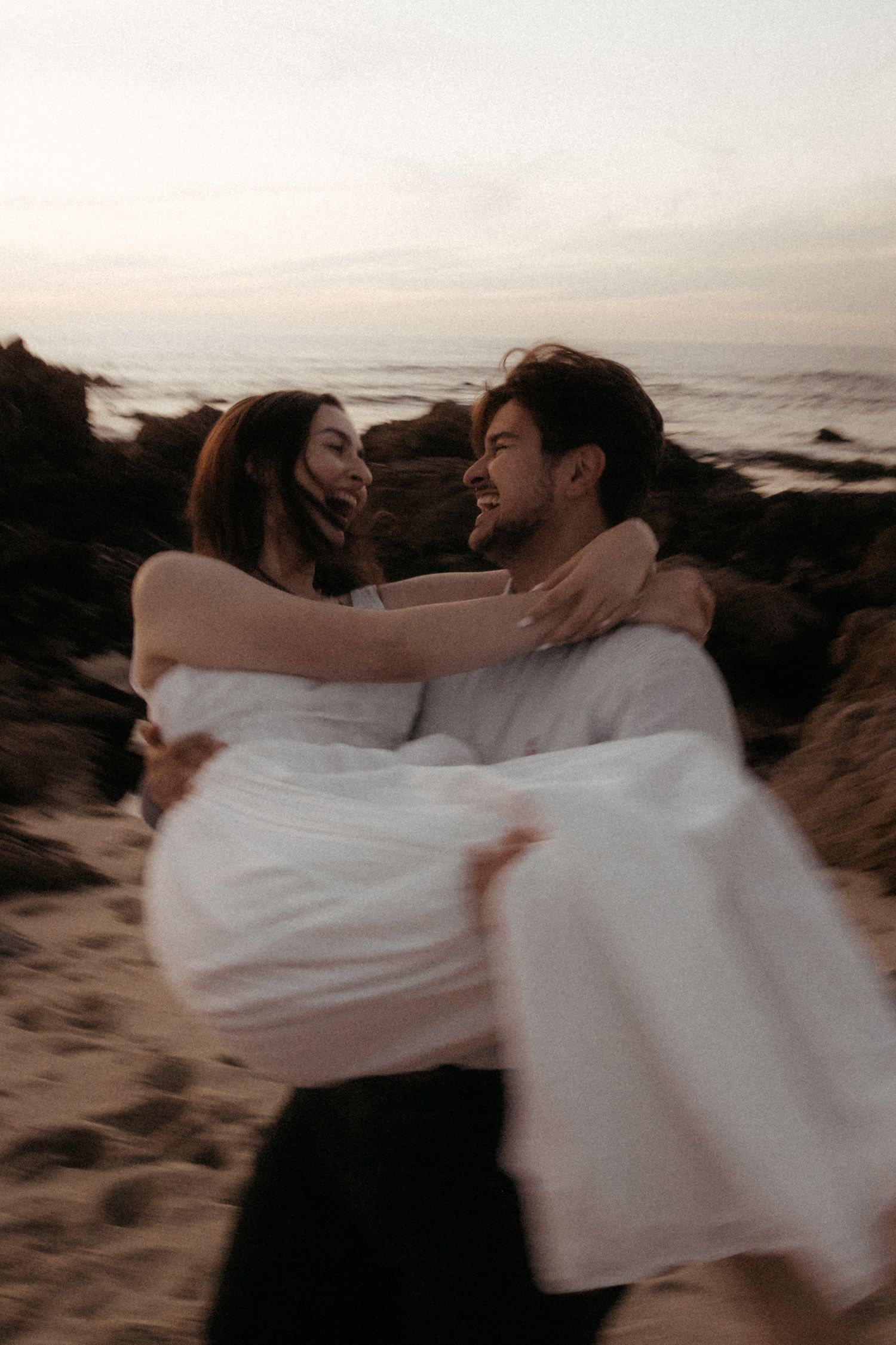 A couple wrapped in white fabric embrace on a sandy beach at sunset creating a romantic silhouette against the ocean view.