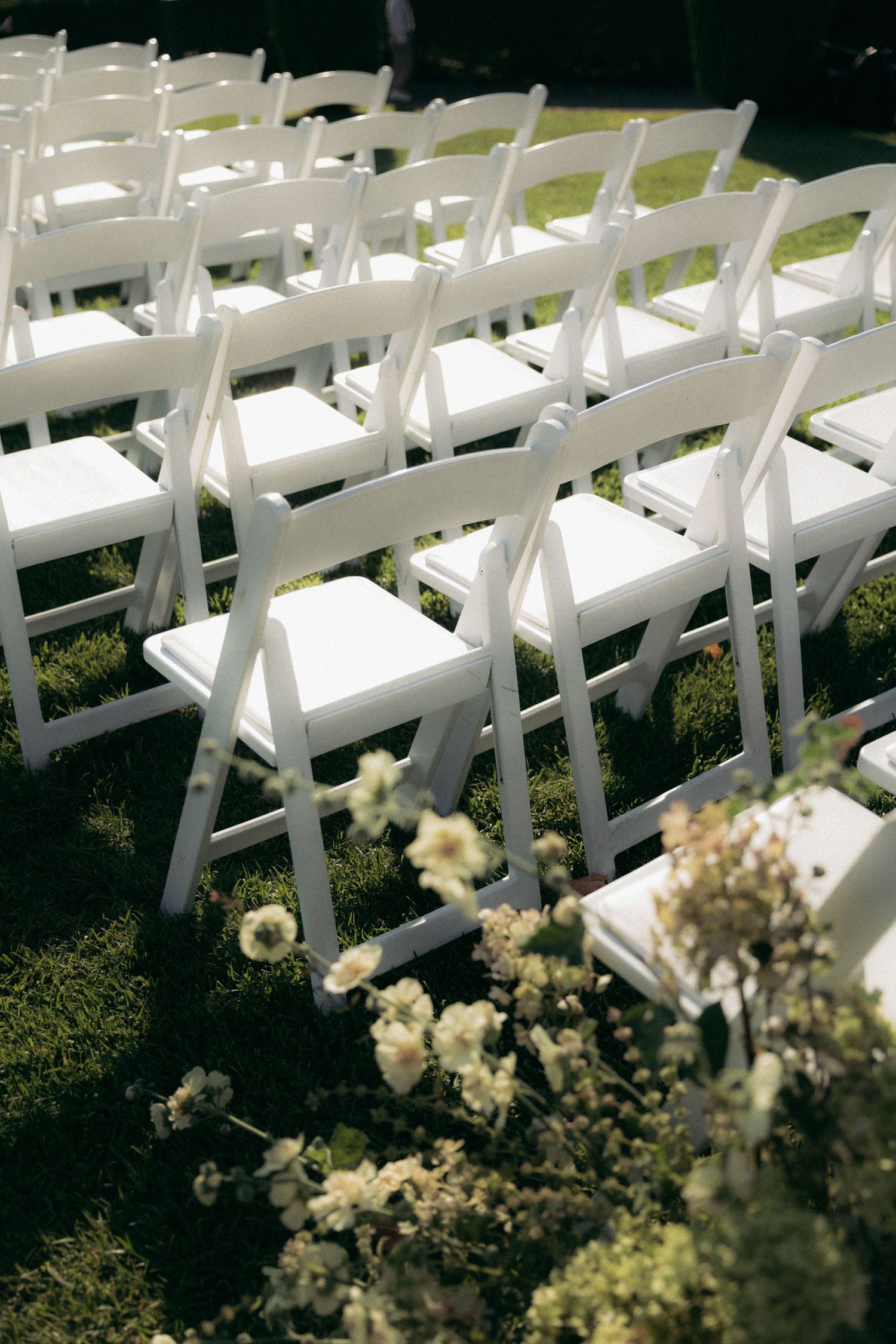 White folding chairs arranged in rows for an outdoor wedding ceremony with delicate floral decorations on the grass.