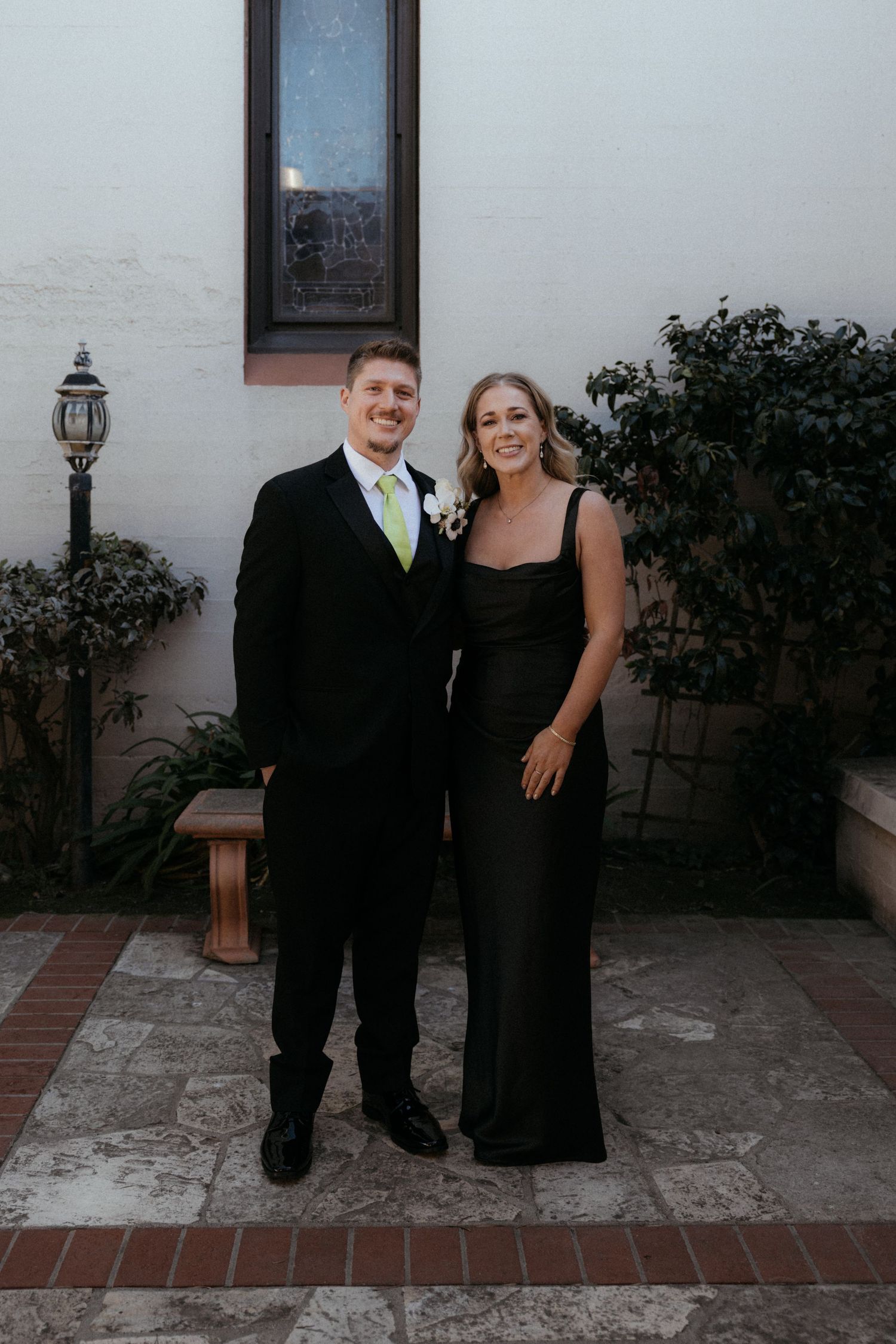 A couple in formal black attire poses together for a series of romantic photos on a paved patio with white walls.