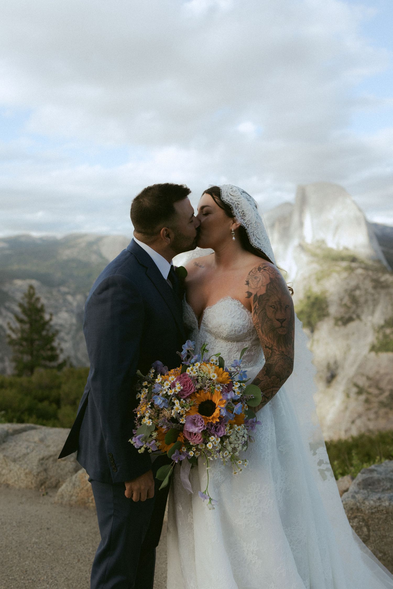 Bride and groom share a kiss at Yosemite overlook, bride holding colorful wildflower bouquet with Half Dome behind them.