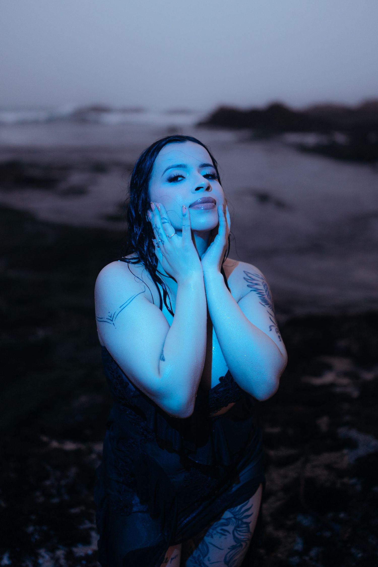 Woman with tattoos poses on rocky beach at dusk, bathed in blue light, hands framing her face.