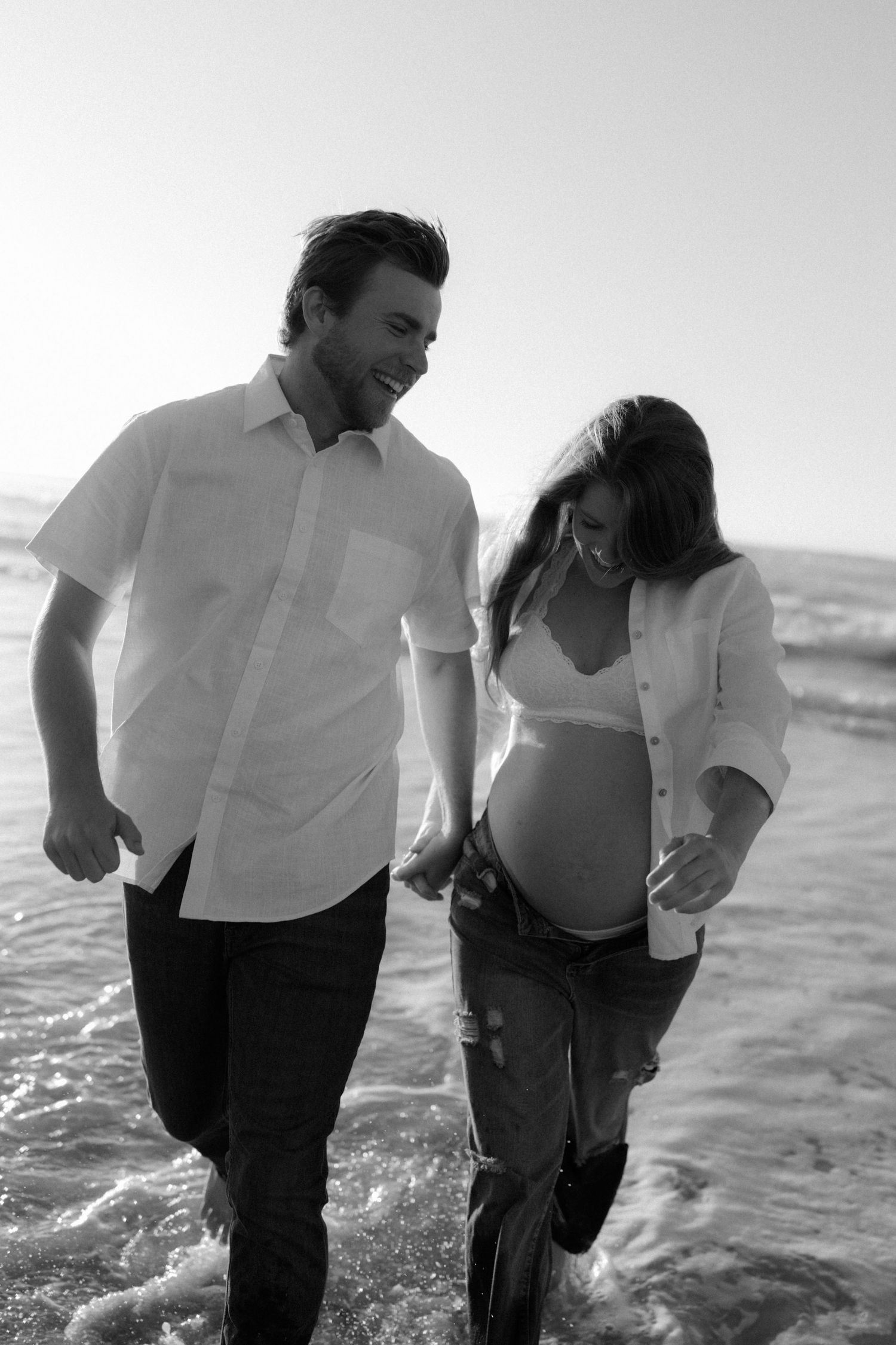 Black and white photo of a happy couple walking hand in hand in ocean waves, woman visibly pregnant.