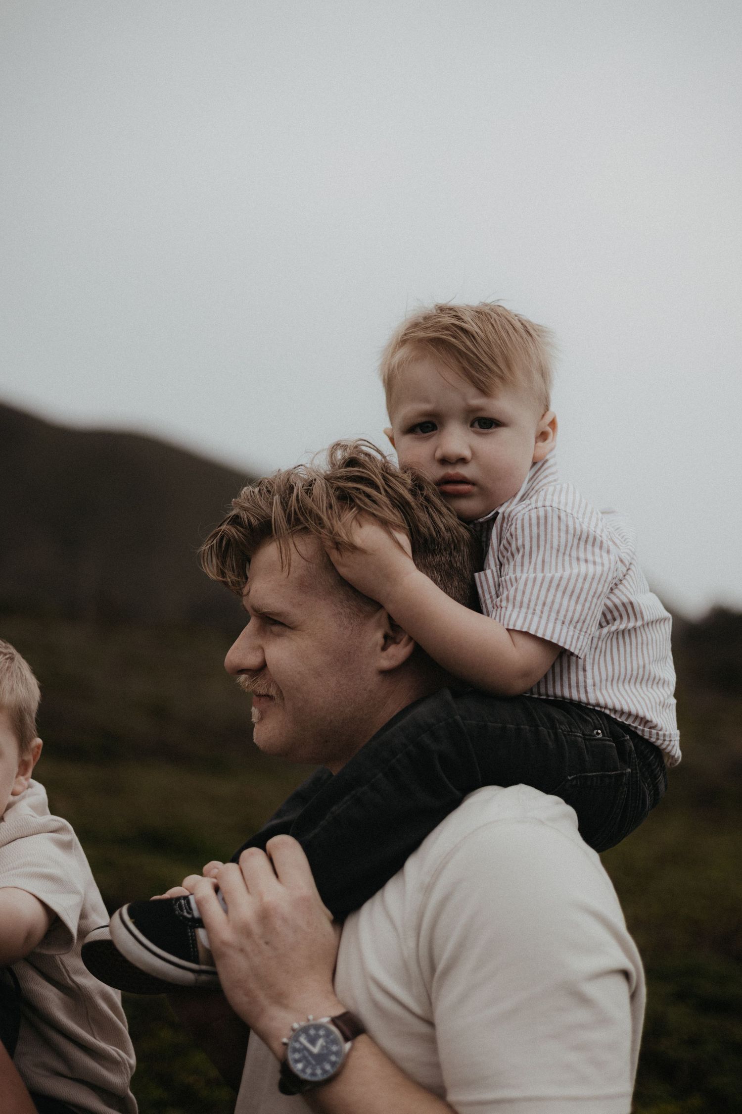 A smiling father carries a young toddler on his shoulders outdoors with green hills in the background.