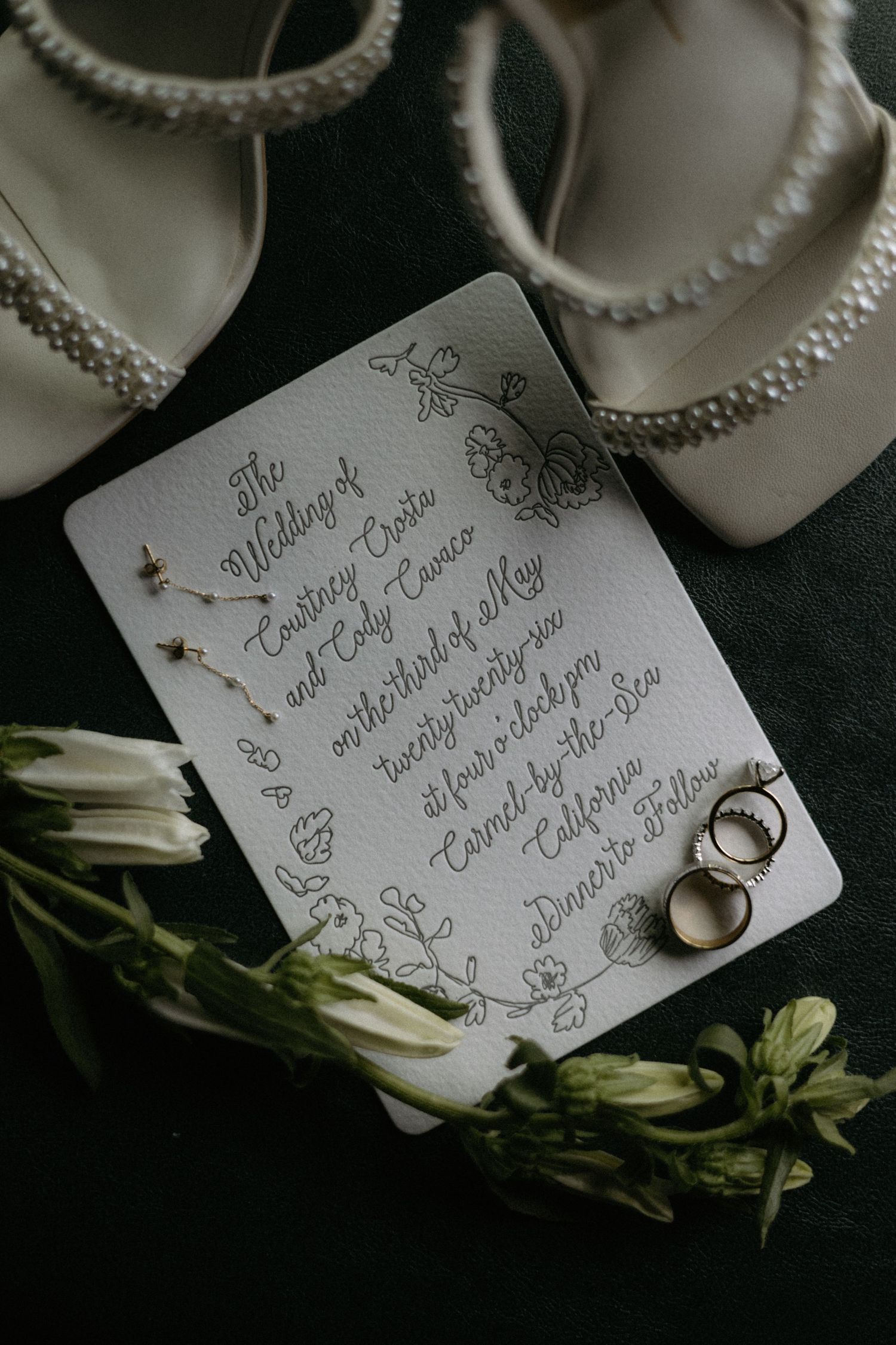 Wedding flat lay featuring invitation, rings, earrings, and pearl-trimmed bridal heels with white florals on dark background.