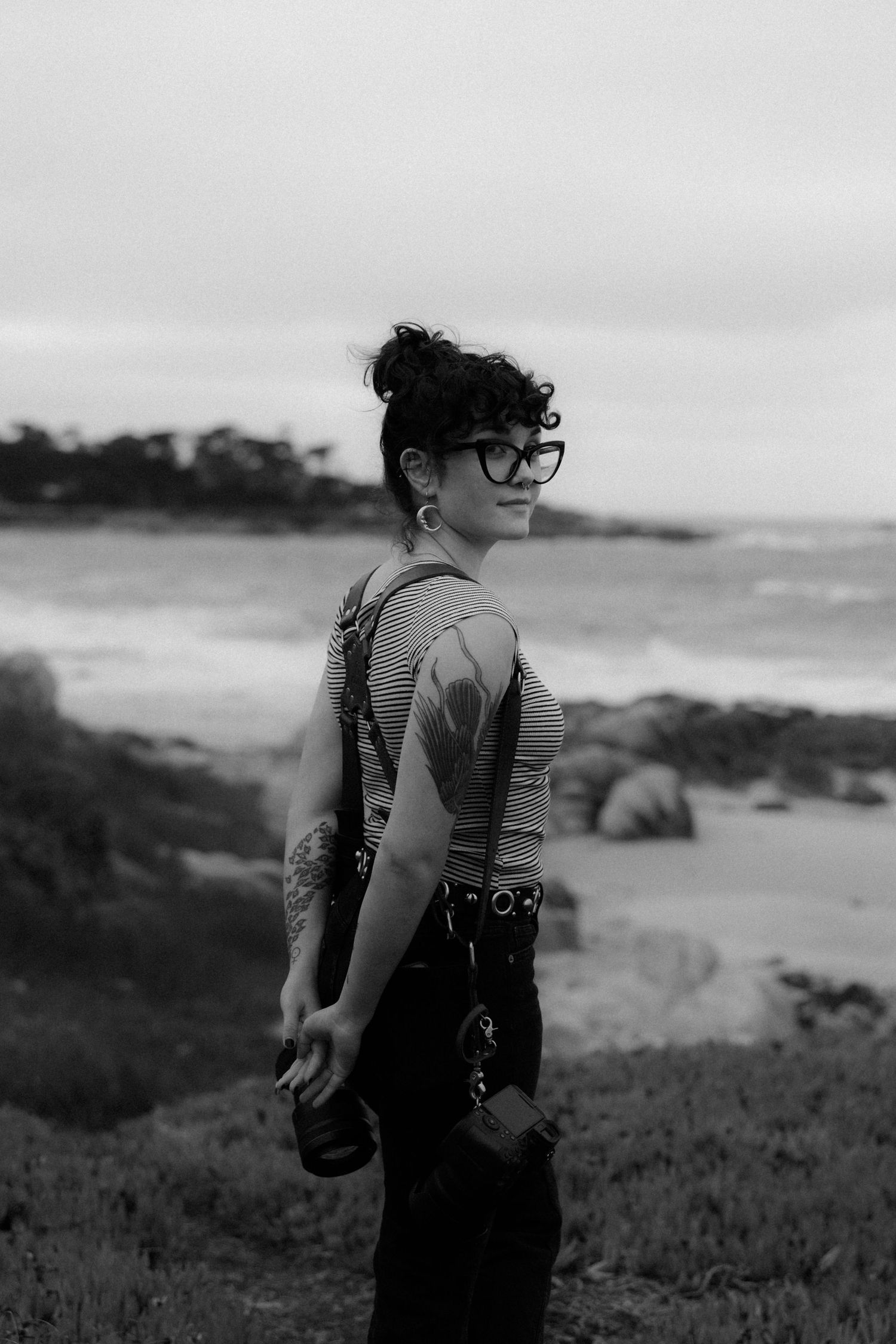 A figure wearing overalls and glasses stands on a hillside overlooking the ocean.