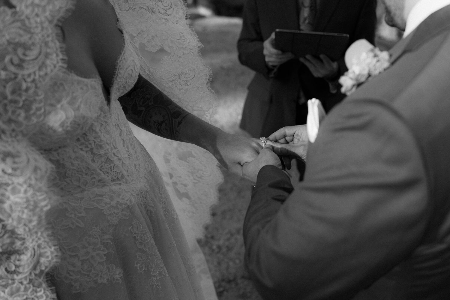 Groom places wedding ring on bride's finger during outdoor ceremony in elegant black and white photo.