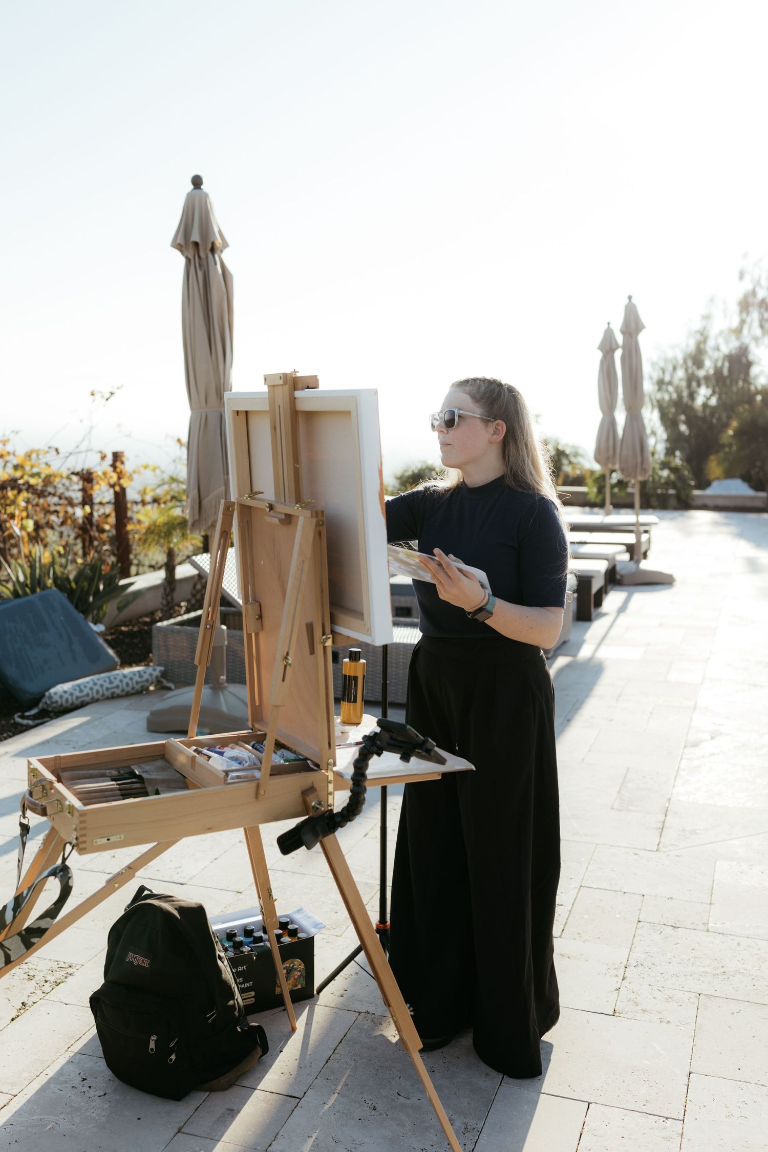 Artist working at an easel on an outdoor terrace with umbrellas and scenic views in the background.