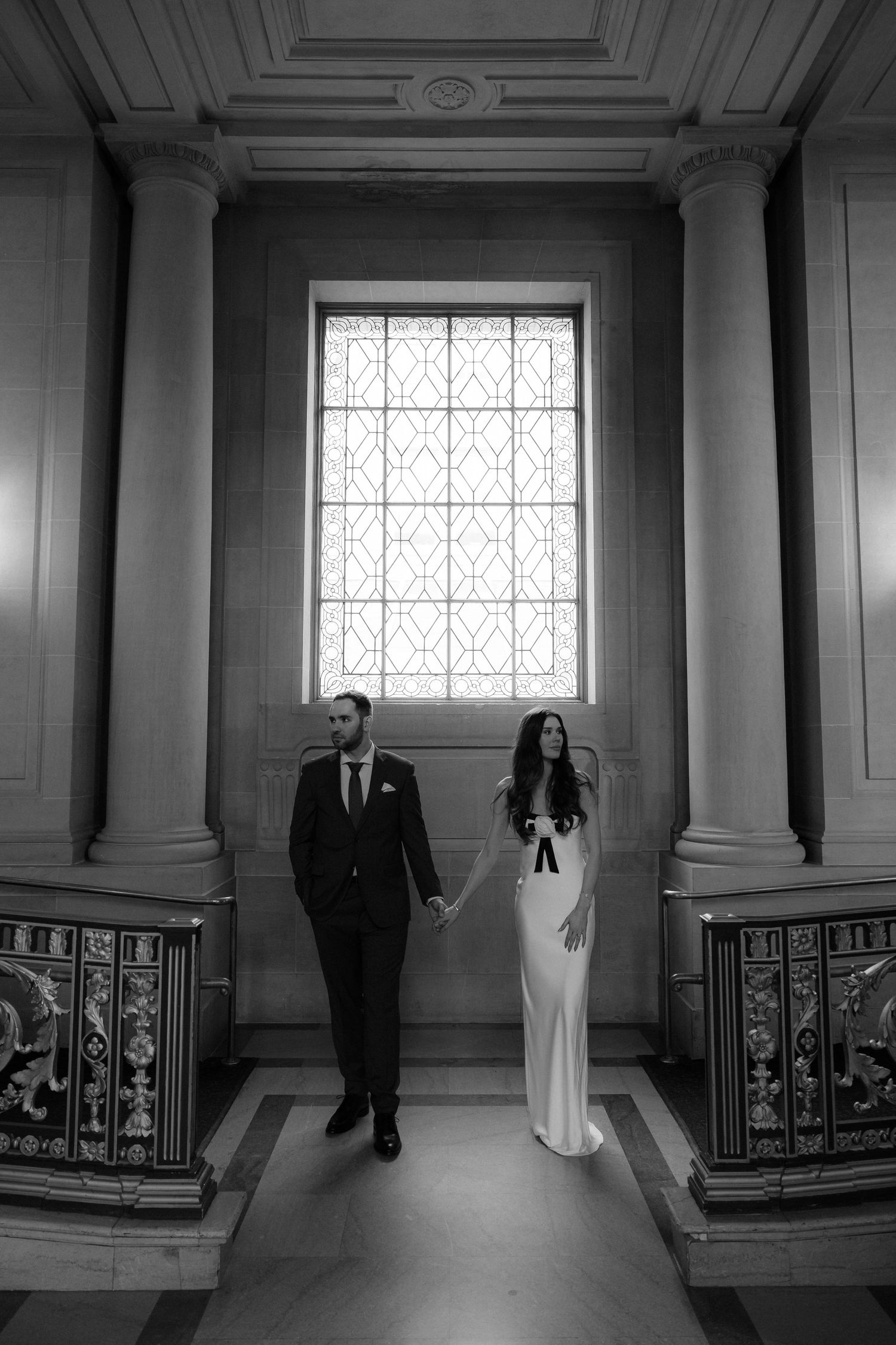Black and white photo of a couple in wedding attire standing in an elegant hallway with columns and a large window.