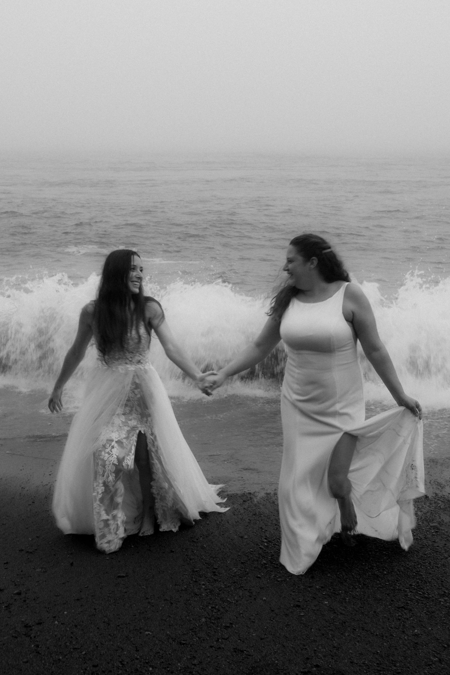 Two people in white dresses hold hands and dance joyfully on a black sand beach with crashing waves behind them.