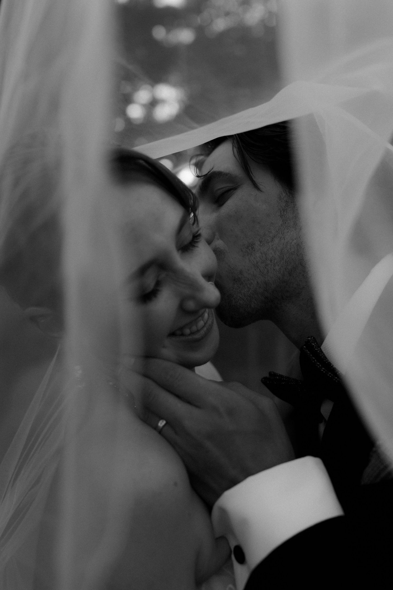 Black and white wedding photo of groom kissing bride on cheek under veil, bride smiling with eyes closed.