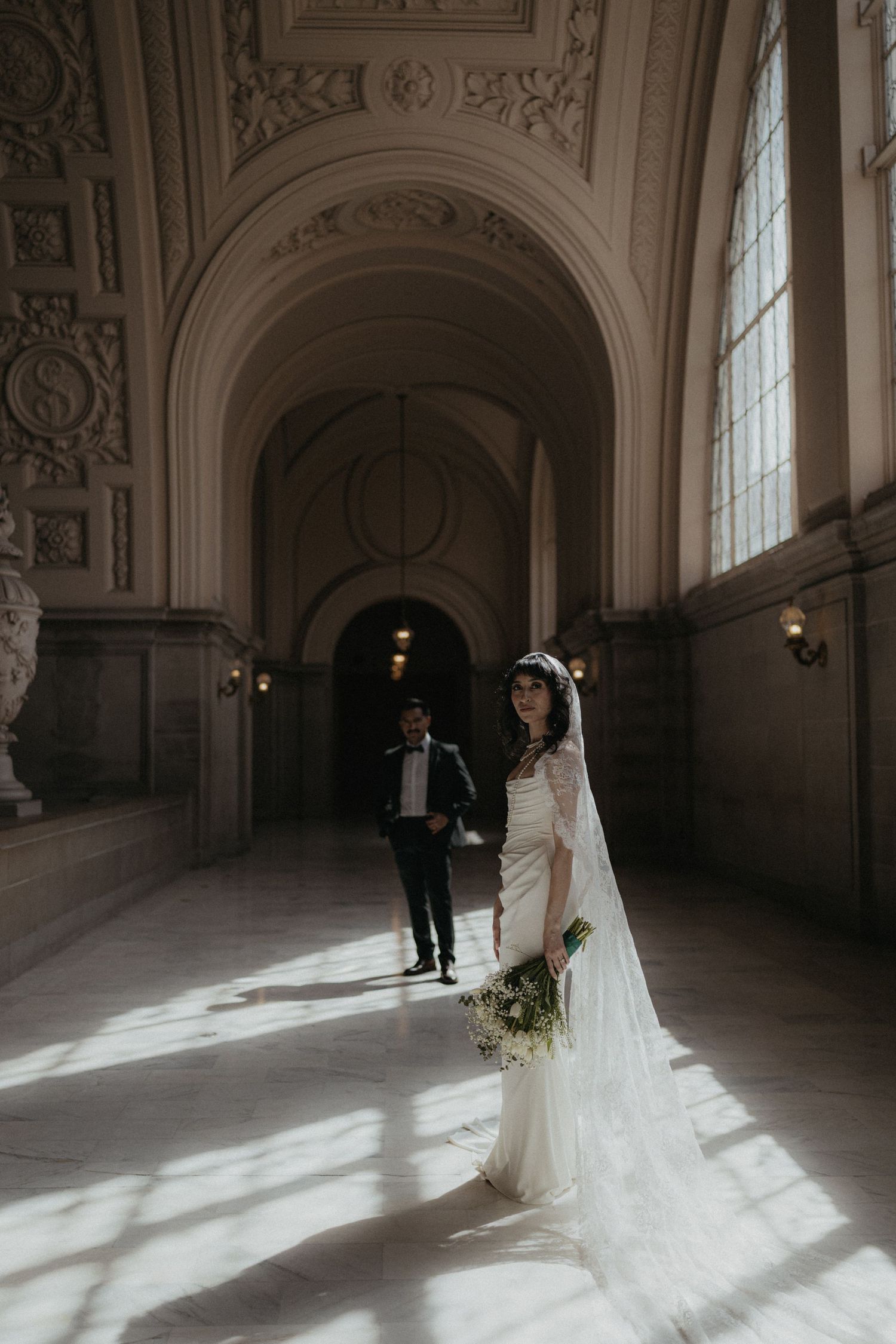 Bride in lace gown with long veil poses in sunlit ornate hallway at San Francisco City Hall.