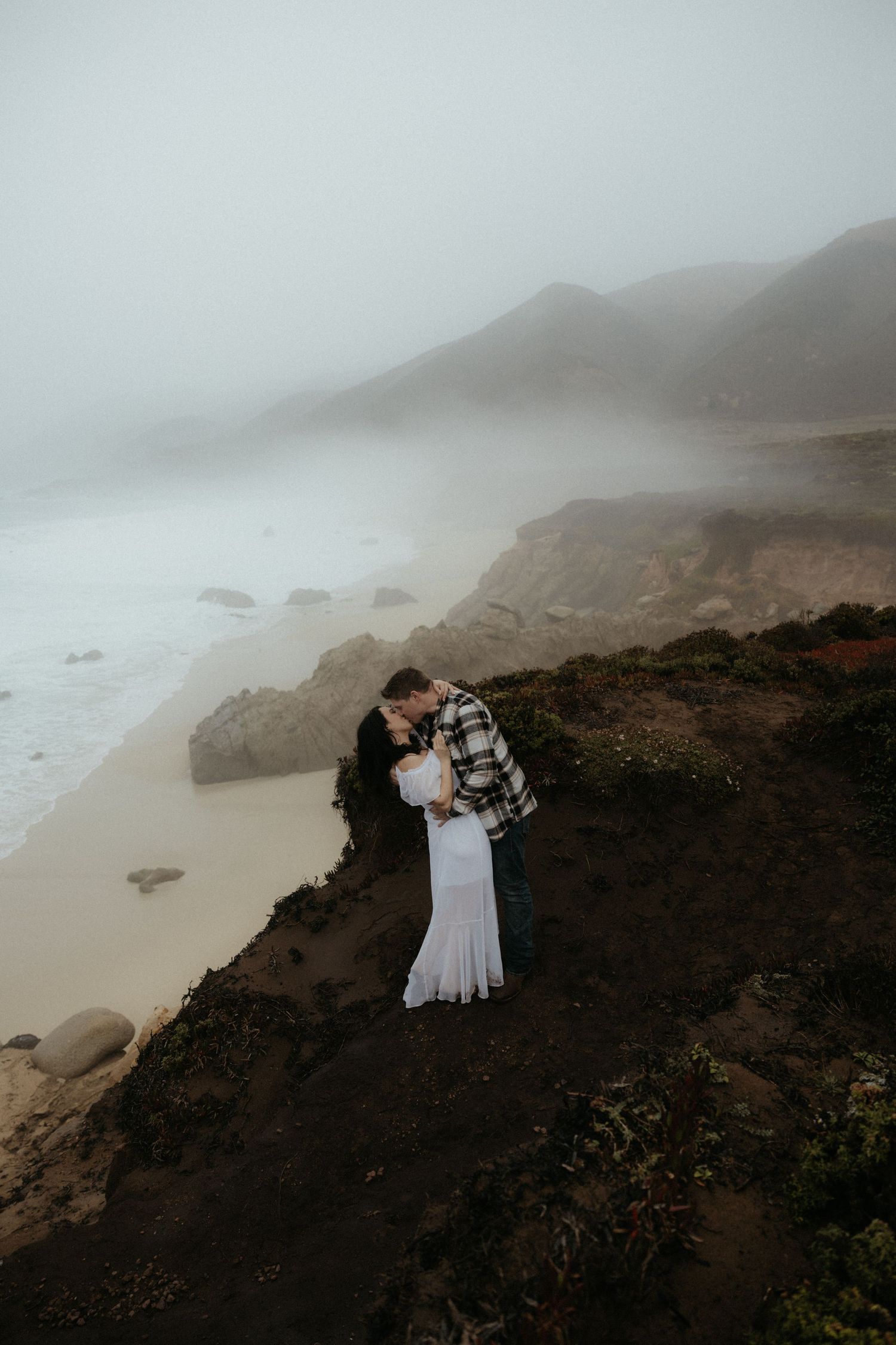 Couple embracing on foggy coastal cliffs overlooking ocean.