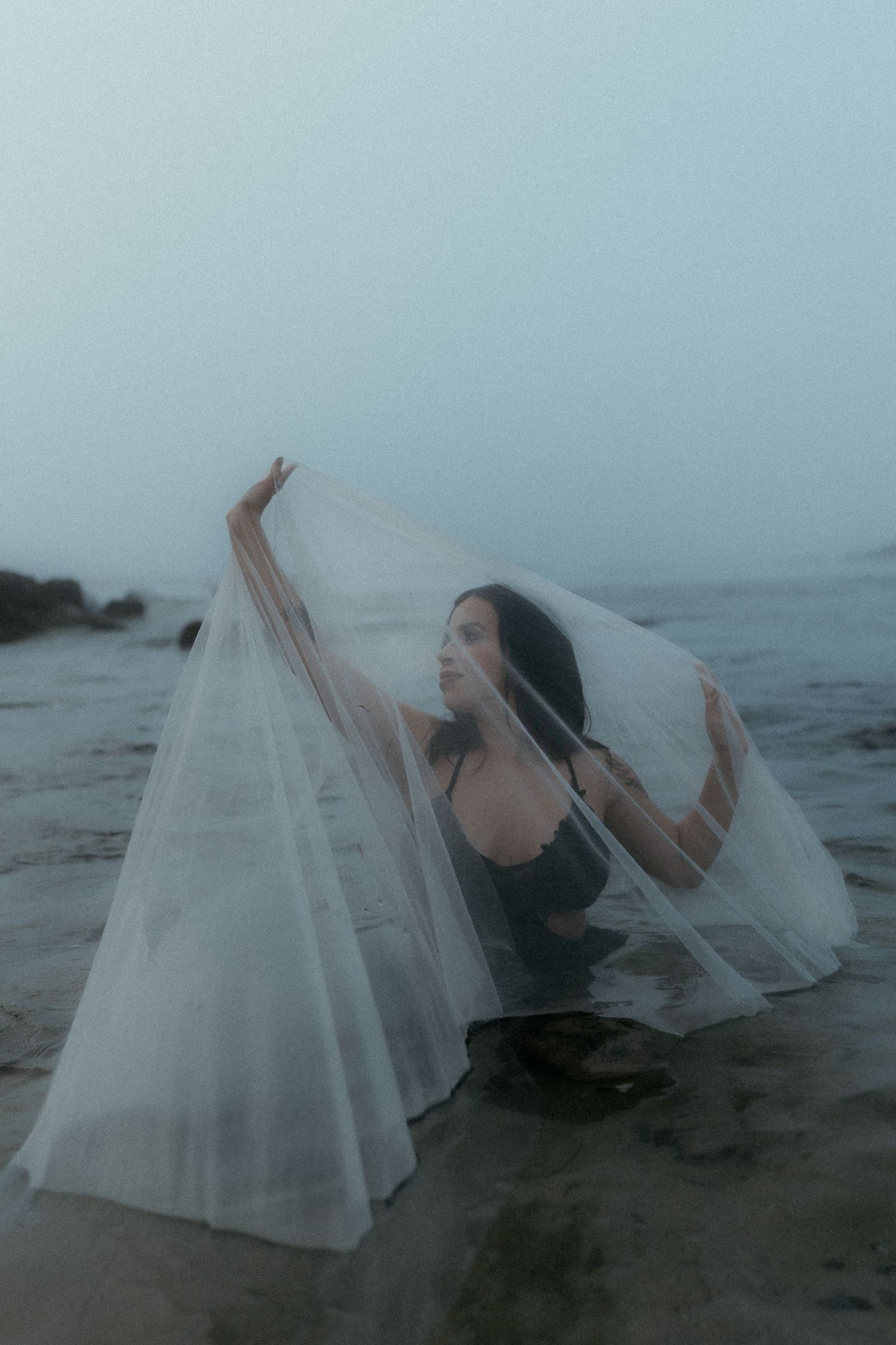 Ethereal figure draped in flowing white veil moves along foggy shoreline.